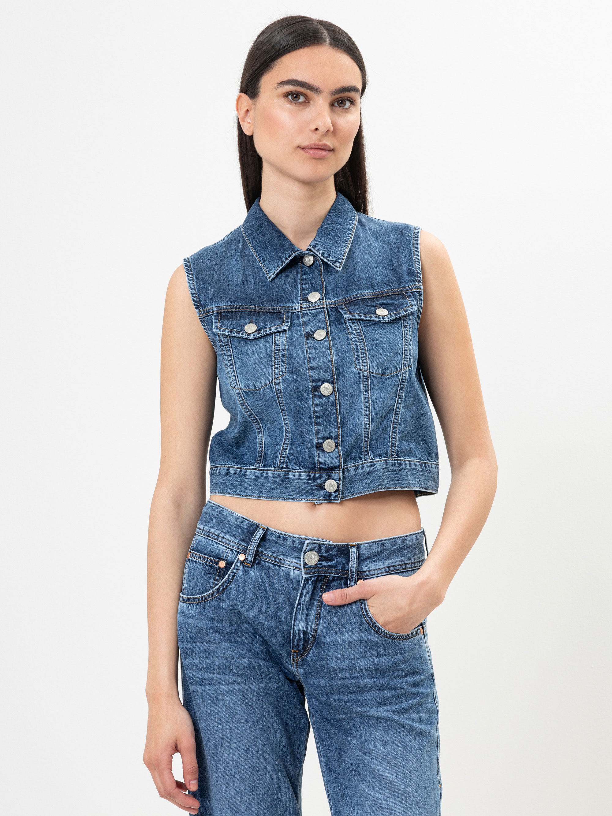 Eine Person mit langen dunklen Haaren trägt eine ärmellose Jeansweste und dazu passende blaue Jeans. Sie steht vor einem schlichten weißen Hintergrund und hat eine Hand in der Tasche.