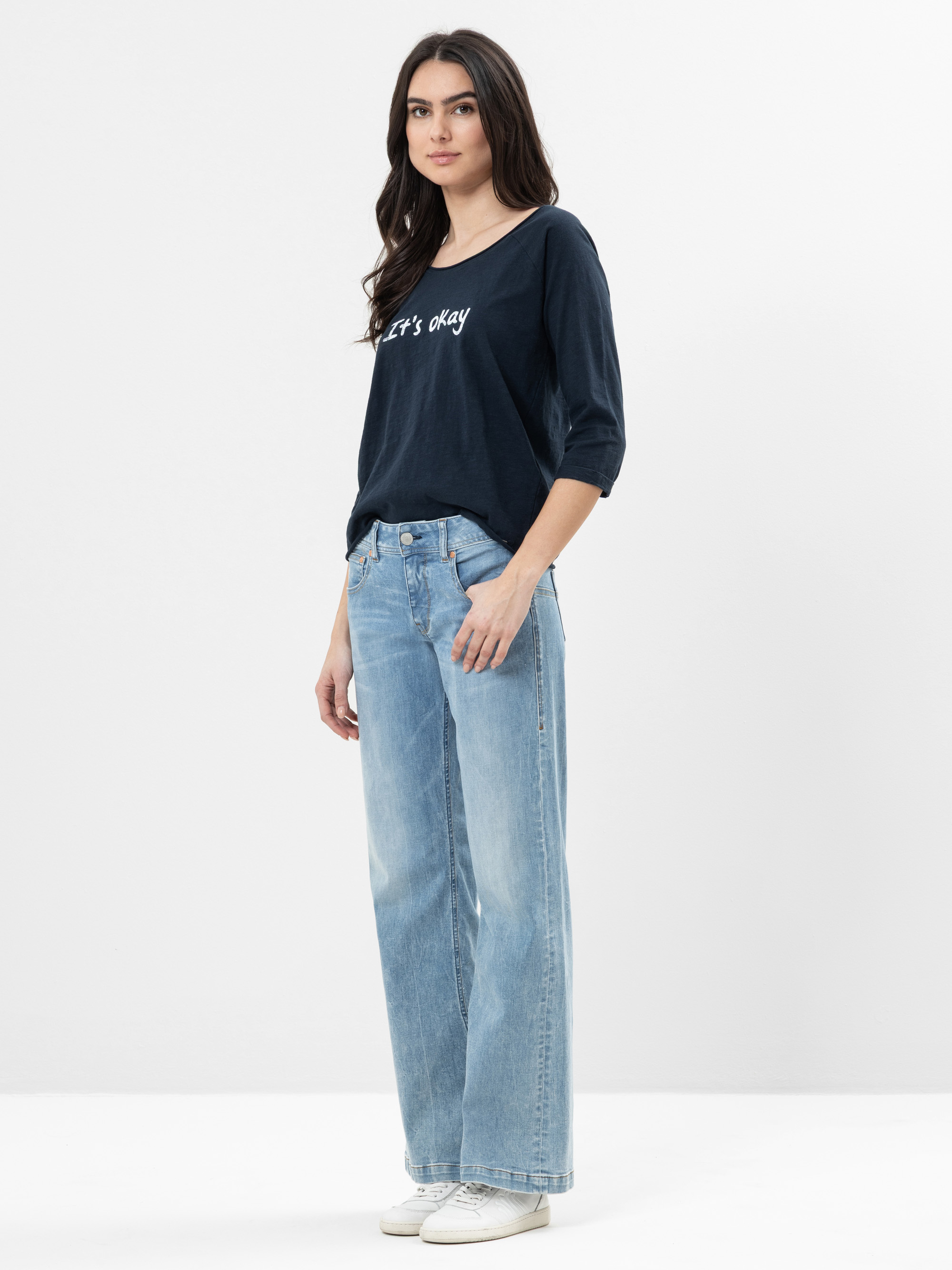 Eine Frau mit langen dunklen Haaren trägt ein marineblaues Hemd mit der Aufschrift "Its okay", hellblaue Jeans mit weitem Bein und weiße Turnschuhe und steht vor einem schlichten weißen Hintergrund.