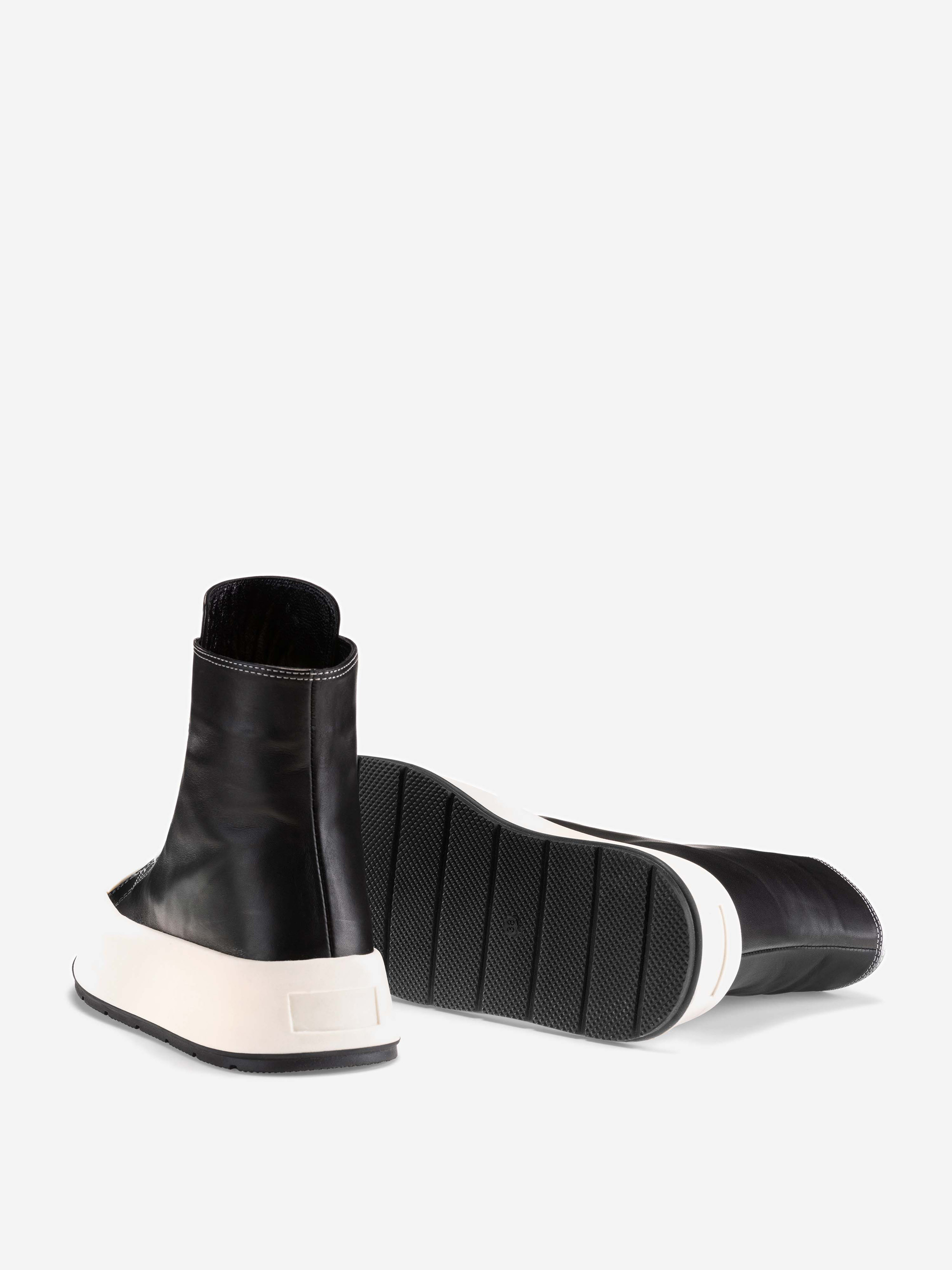 Ein Paar schwarzer High-Top-Sneakers mit dicken weißen Sohlen; ein Schuh steht aufrecht, während der andere auf der Seite liegt und die strukturierte Sohle zeigt. Der Hintergrund ist hellgrau.