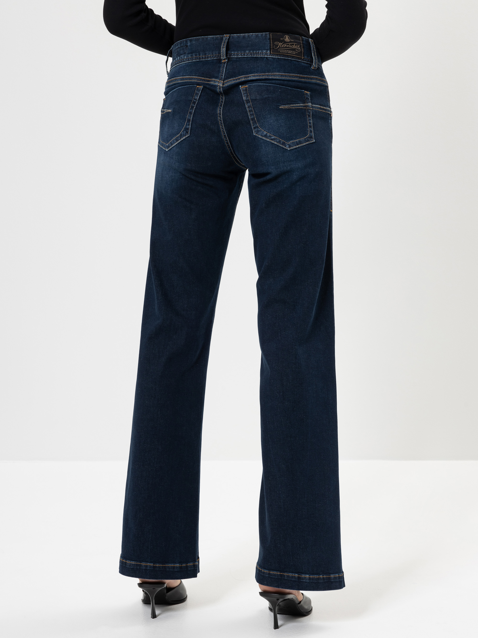 Eine Person, die dunkelblaue Jeans mit geradem Bein und schwarze Schuhe mit Absätzen trägt, steht mit dem Gesicht nach außen, die Hände in die Hüften gestützt, vor einem schlichten weißen Hintergrund.