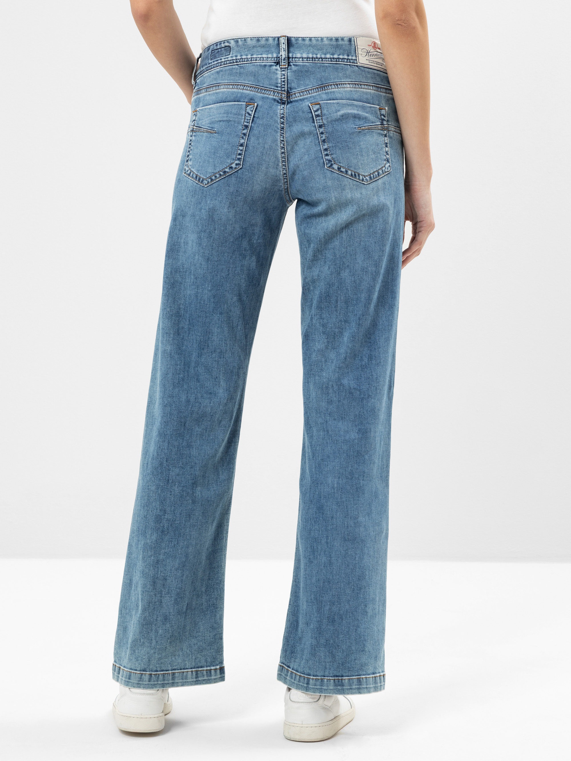 Eine Person, die hoch taillierte, weit geschnittene Bluejeans und weiße Turnschuhe trägt, steht mit dem Rücken zur Kamera vor einem schlichten weißen Hintergrund.