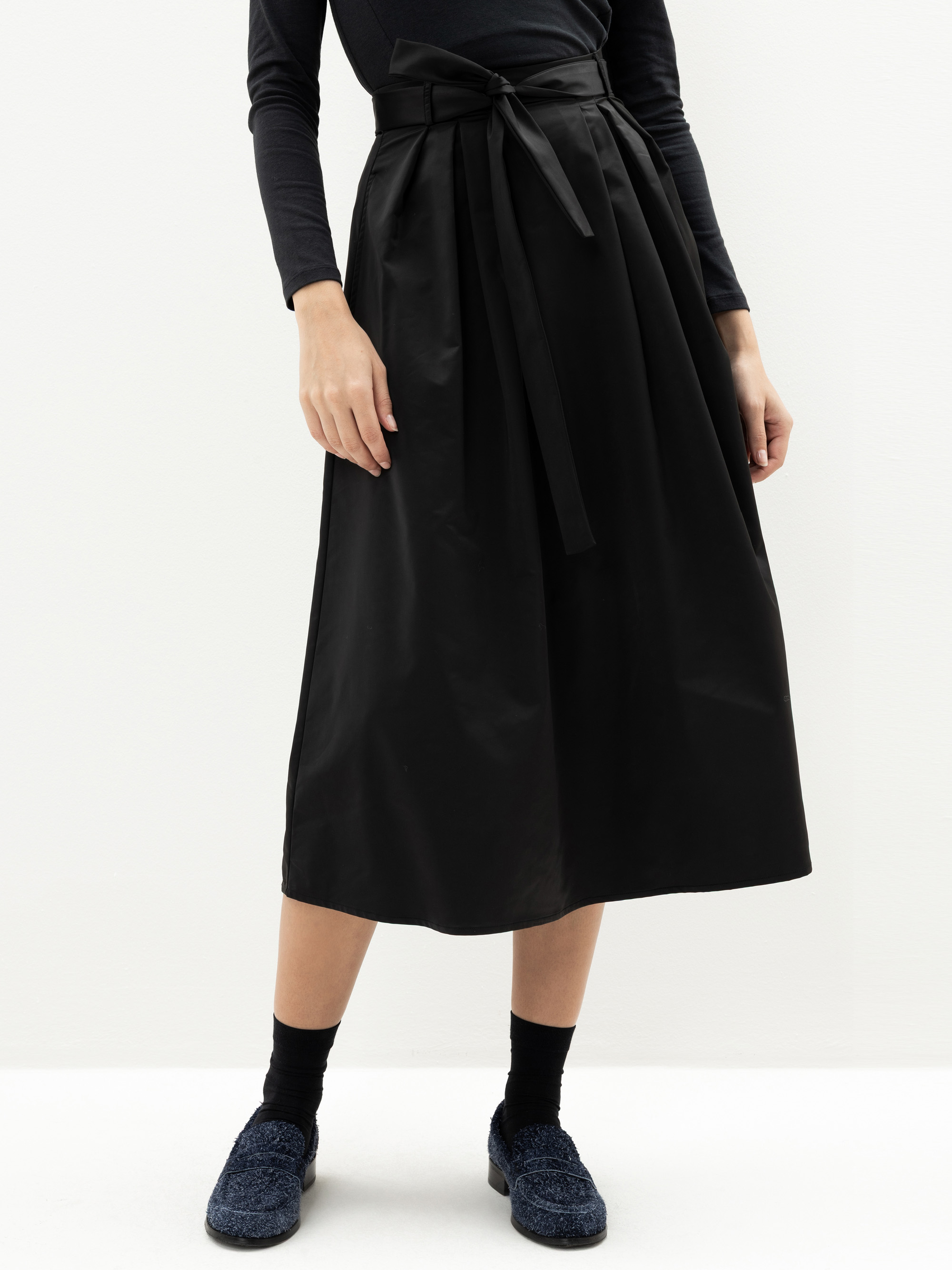 Eine Person, die ein schwarzes langärmeliges Oberteil, einen hochtaillierten schwarzen Midirock mit Bindegürtel, schwarze Socken und marineblaue, strukturierte Schuhe trägt, steht vor einem schlichten weißen Hintergrund.