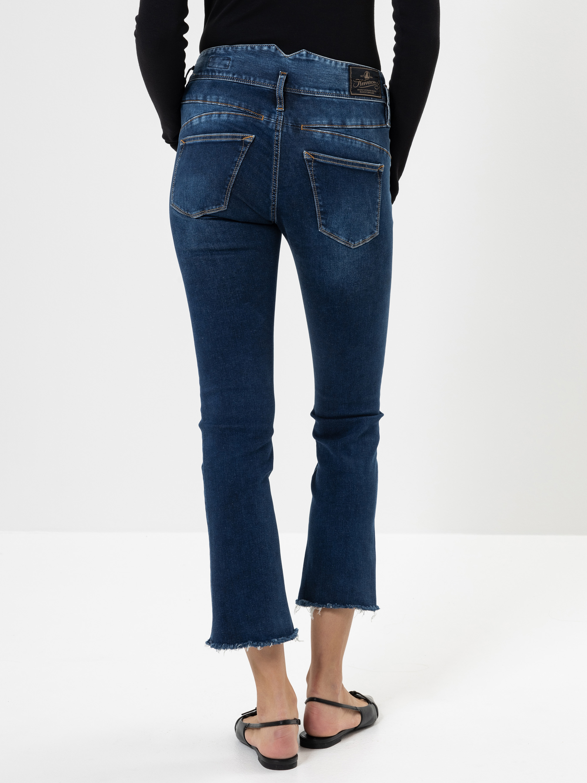Eine Person, die dunkelblaue, leicht abgeschnittene Jeans mit ausgefransten Säumen und schwarze Sandalen trägt, steht mit dem Gesicht zur Kamera vor einem schlichten weißen Hintergrund.