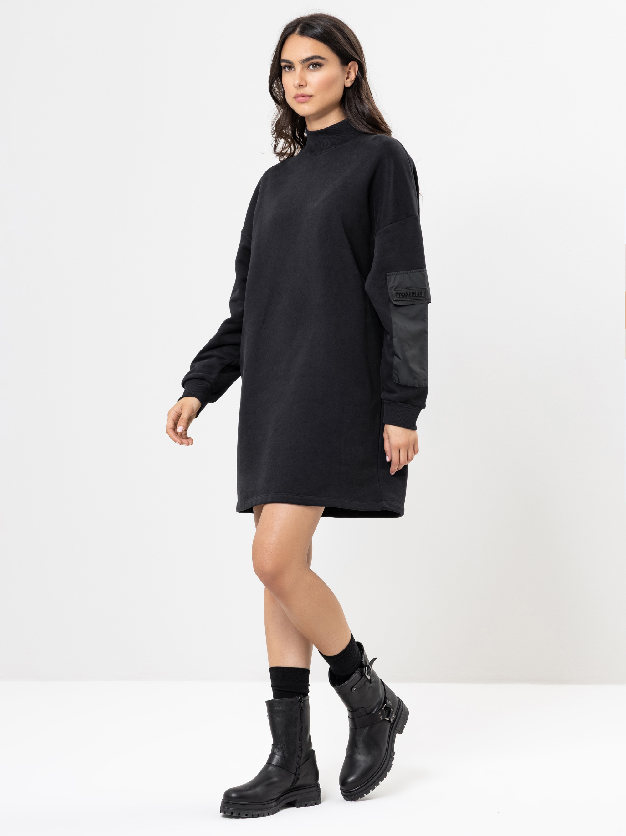 Eine Frau mit langen dunklen Haaren trägt ein lockeres schwarzes Pulloverkleid mit langen Ärmeln, schwarze Socken und schwarze Kampfstiefel und steht vor einem schlichten weißen Hintergrund.