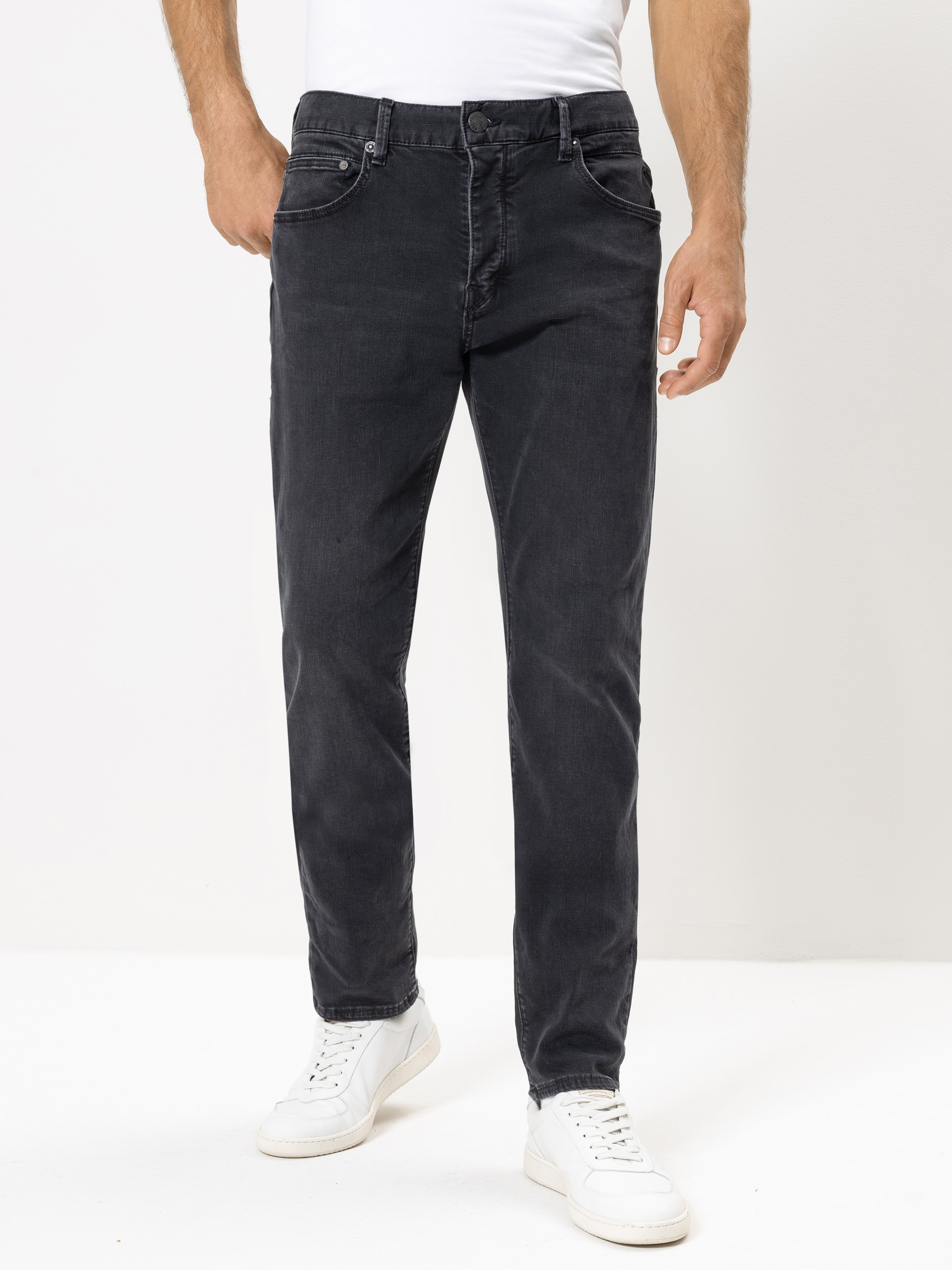 Ein Mann, der ein weißes T-Shirt, dunkelgraue Jeans und weiße Turnschuhe trägt, steht vor einem schlichten weißen Hintergrund. Das Foto ist von den Schultern abwärts abgeschnitten und konzentriert sich auf die Jeans.