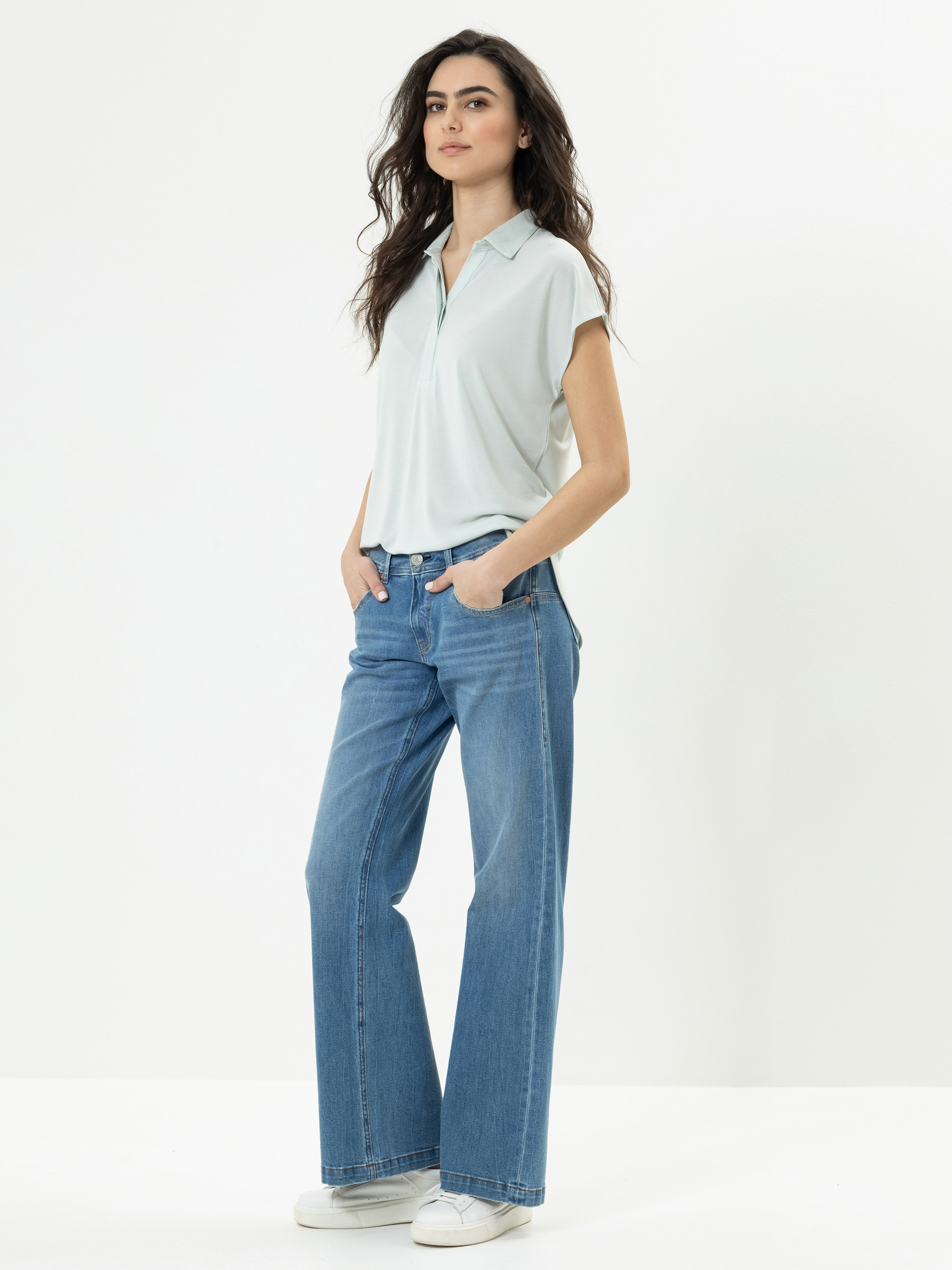 Eine Frau mit langen dunklen Haaren steht vor einem schlichten weißen Hintergrund. Sie trägt ein hellblaues, kurzärmeliges Poloshirt, blaue, weit geschnittene Jeans und weiße Turnschuhe. Sie hat die Hände in den Taschen und einen entspannten Gesichts