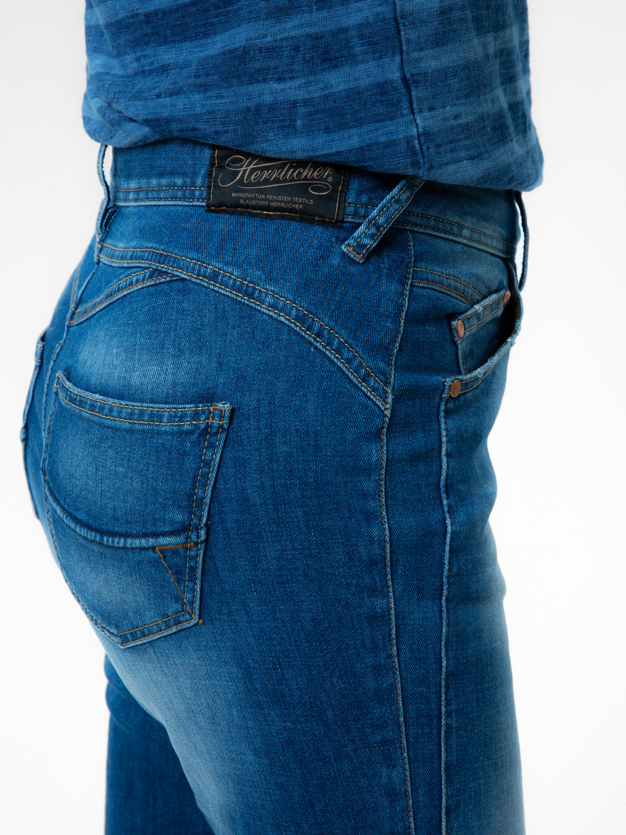 Person, die eine blaue Jeans und ein blaues Hemd trägt, dargestellt von der Taille abwärts mit Schwerpunkt auf der Gesäßtasche und dem schwarzen Markenetikett der Jeans am Bund. Der Hintergrund ist einfarbig und weiß.