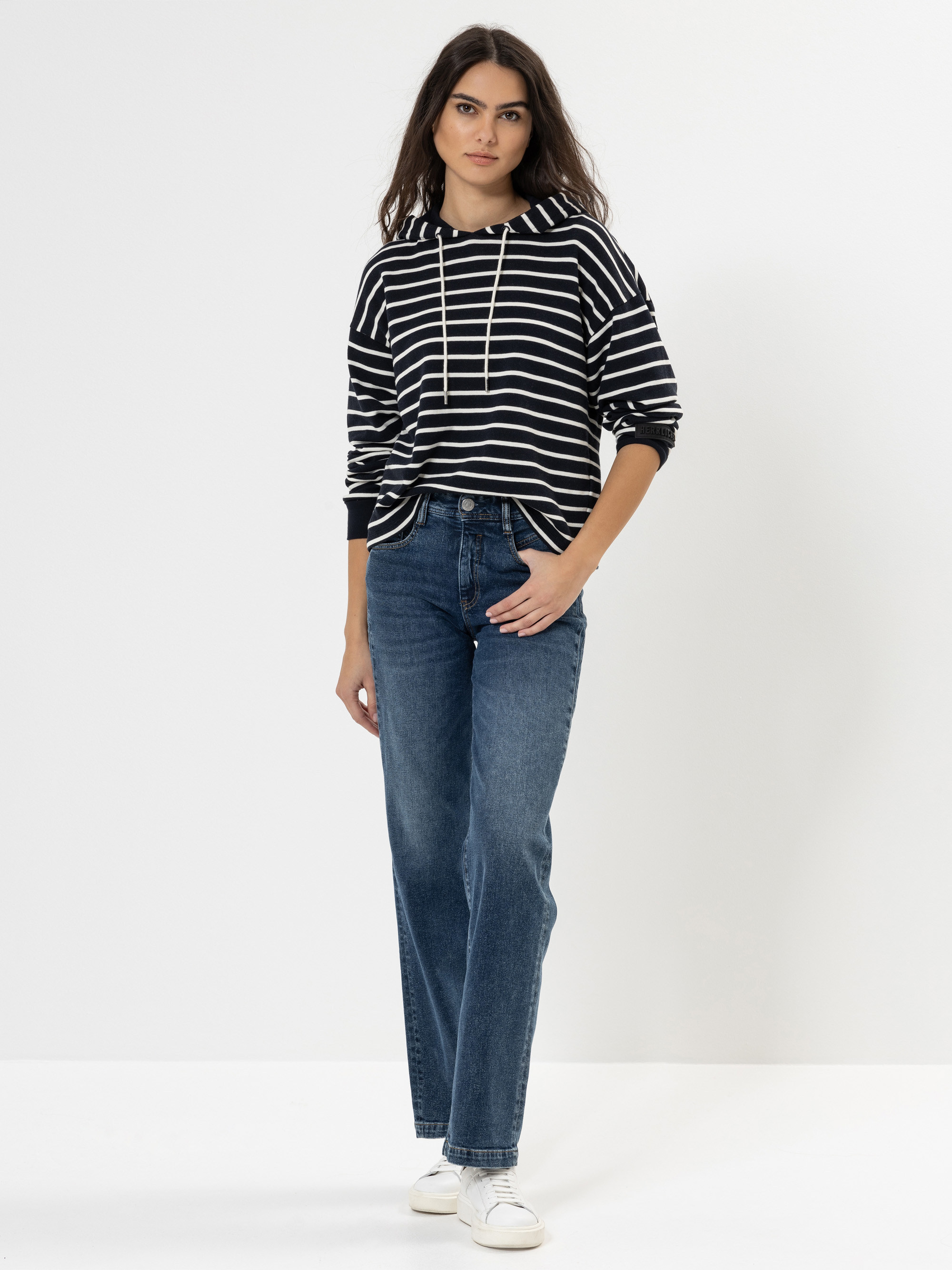 Eine Frau steht vor einem schlichten weißen Hintergrund und trägt einen schwarz-weiß gestreiften Kapuzenpullover, blaue Jeans und weiße Turnschuhe. Sie hat langes dunkles Haar und einen neutralen Gesichtsausdruck, eine Hand steckt in ihrer Tasche.