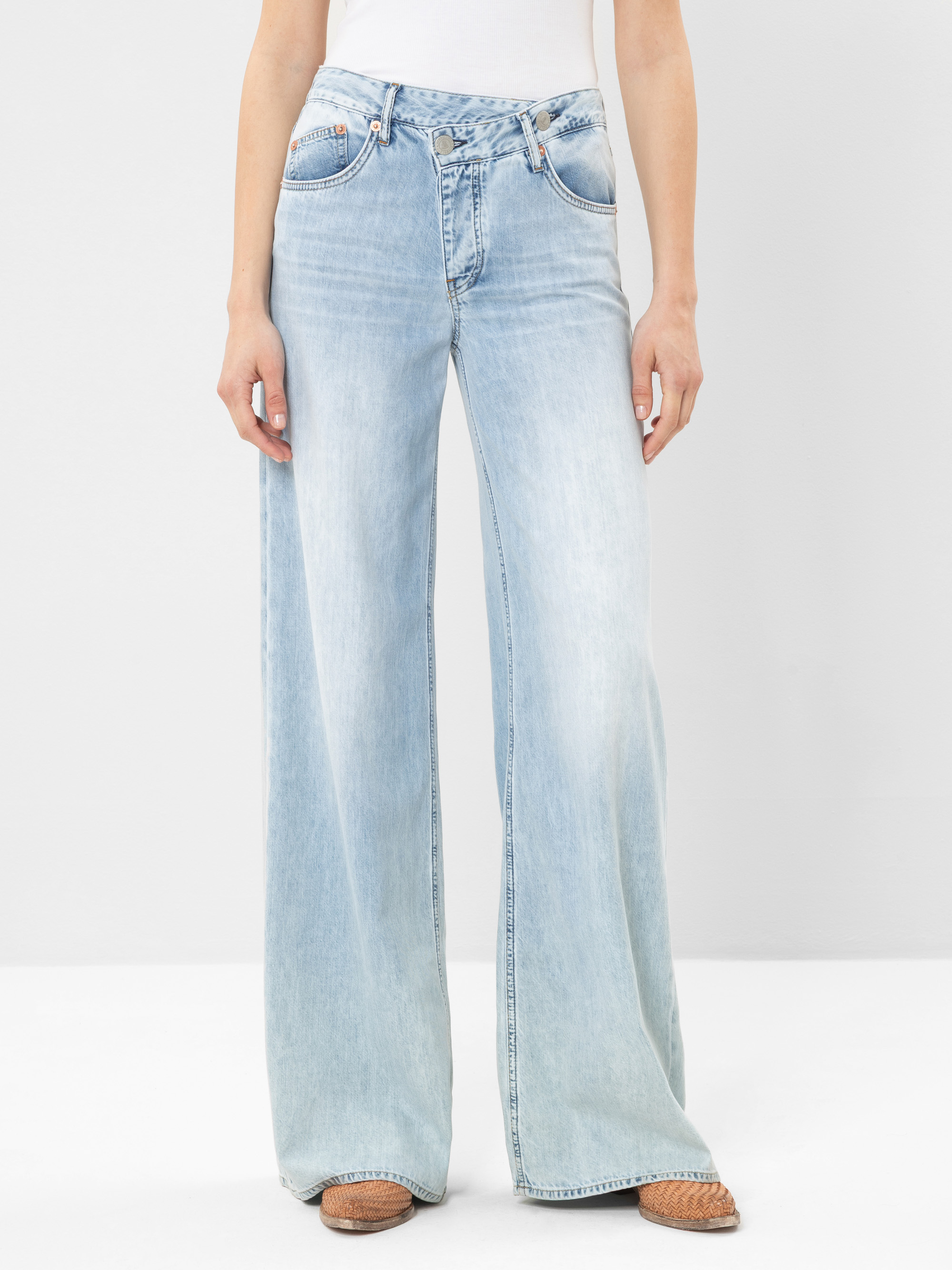 Eine Person, die hellblaue Jeans mit hoher Taille und weitem Bein, ein weißes Oberteil und braune gewebte Schuhe trägt, steht vor einem schlichten weißen Hintergrund.