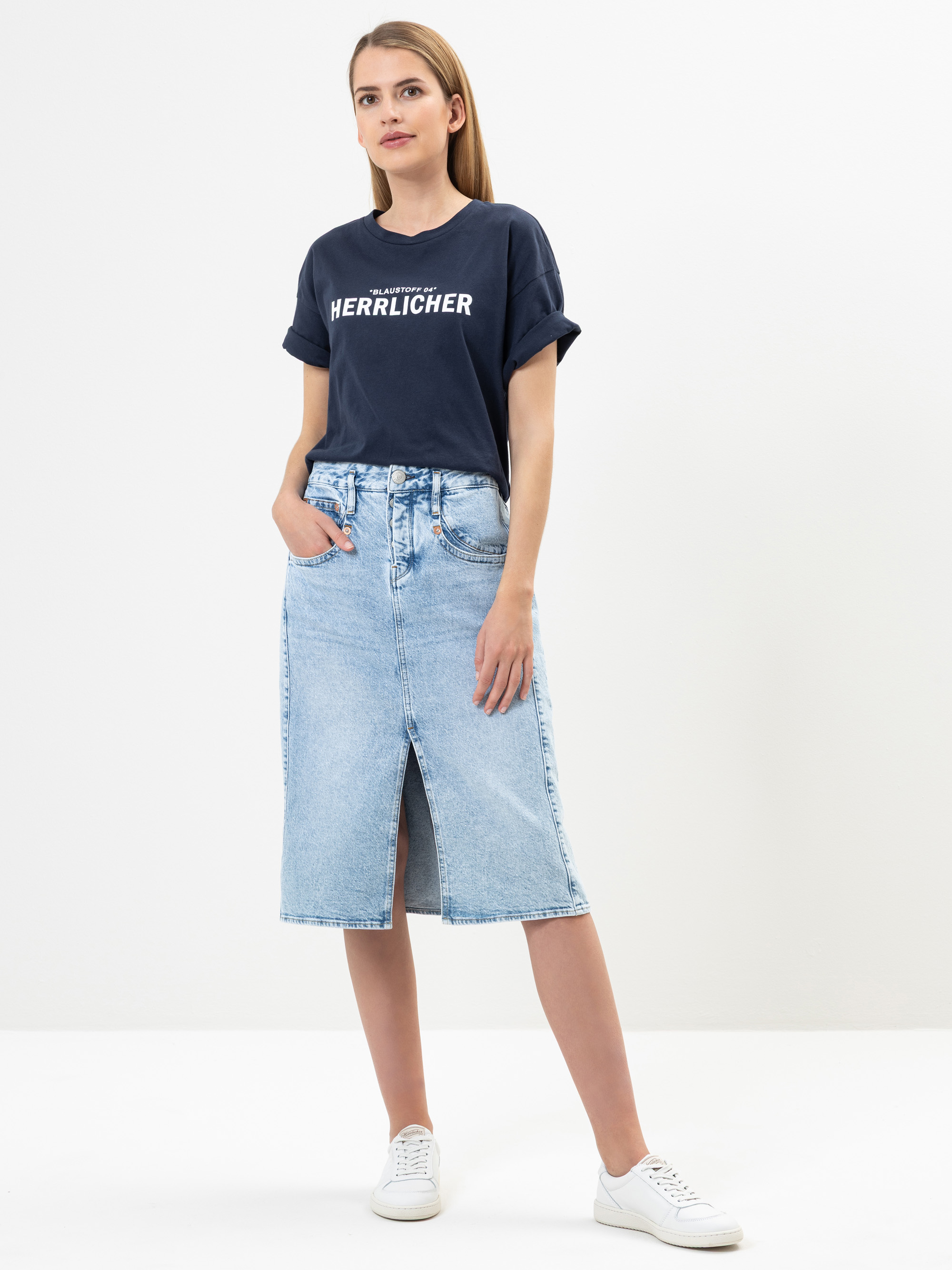 Eine Frau steht vor einem weißen Hintergrund und trägt ein marineblaues Herrlicher-T-Shirt, einen hellblauen Jeans-Midirock mit einem Schlitz vorne und weiße Turnschuhe. Sie hat die Hände in den Taschen und blickt selbstbewusst nach vorne.