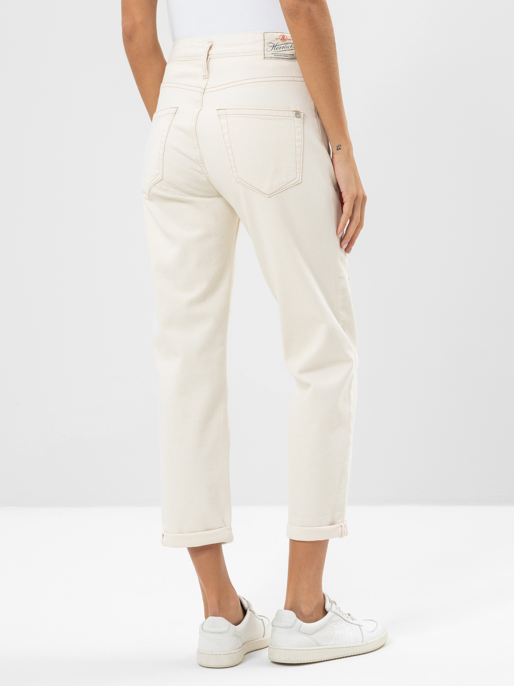 Eine Person steht mit dem Gesicht zur Wand und trägt eine cremefarbene Jeans mit Bündchen, ein weißes Oberteil und weiße Turnschuhe vor einem schlichten hellen Hintergrund. Die Jeans hat Gesäßtaschen und einen sichtbaren Markenaufnäher am Bund.