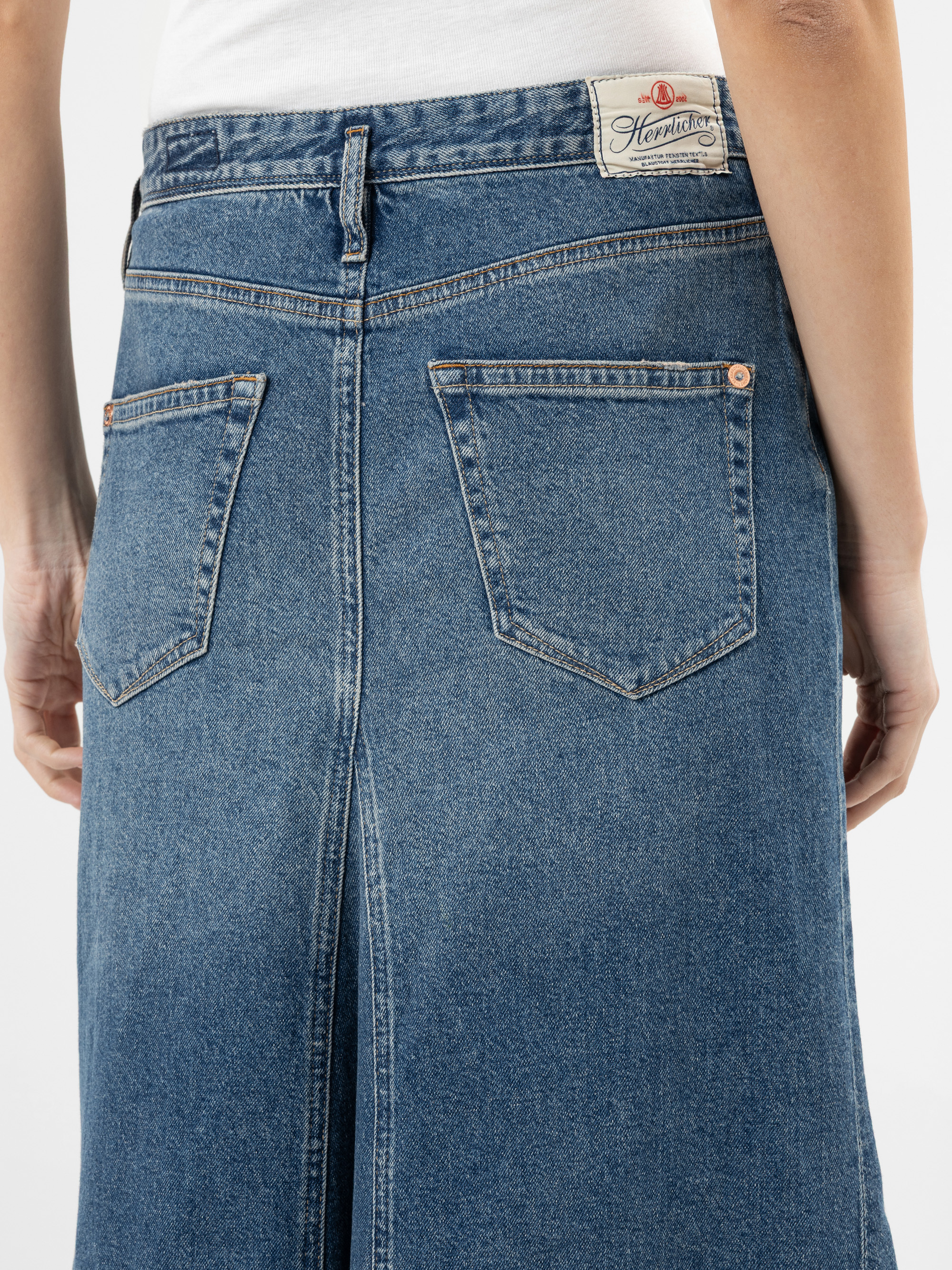 Eine Person, die einen blauen Jeansrock mit Gesäßtaschen und einem weißen Etikett am Bund trägt, steht vor einem schlichten Hintergrund.