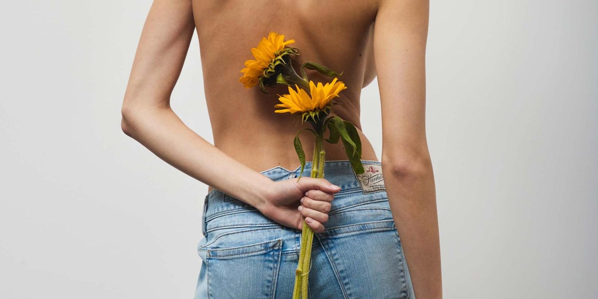Eine Person mit nacktem Rücken und in blauen Jeans hält zwei Sonnenblumen hinter ihrem Rücken vor einem schlichten, hellen Hintergrund.