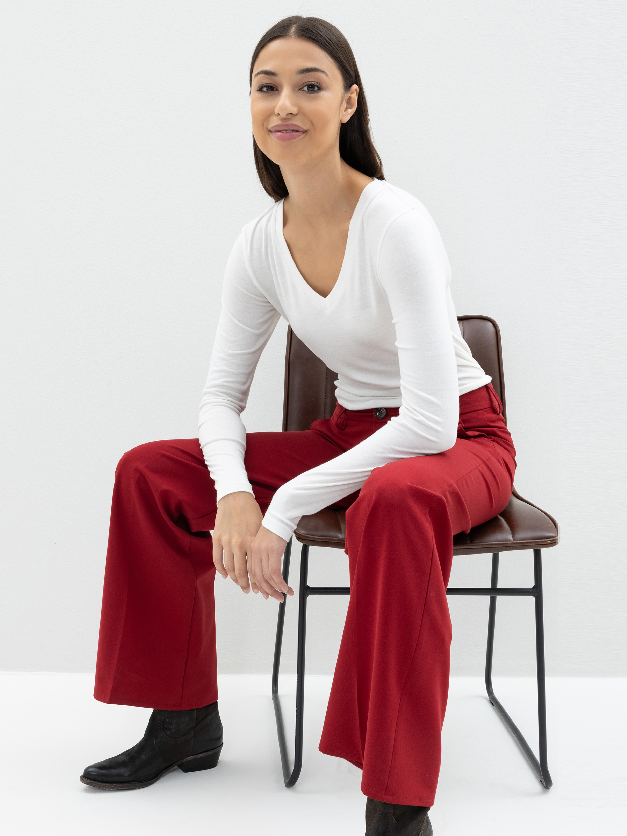 Eine Frau mit glattem dunklem Haar, die ein weißes langärmeliges Oberteil und eine rote Hose mit weitem Bein trägt, sitzt auf einem braunen Stuhl vor einem schlichten weißen Hintergrund und lächelt sanft, wobei sie einen Arm auf ihr Knie stützt.