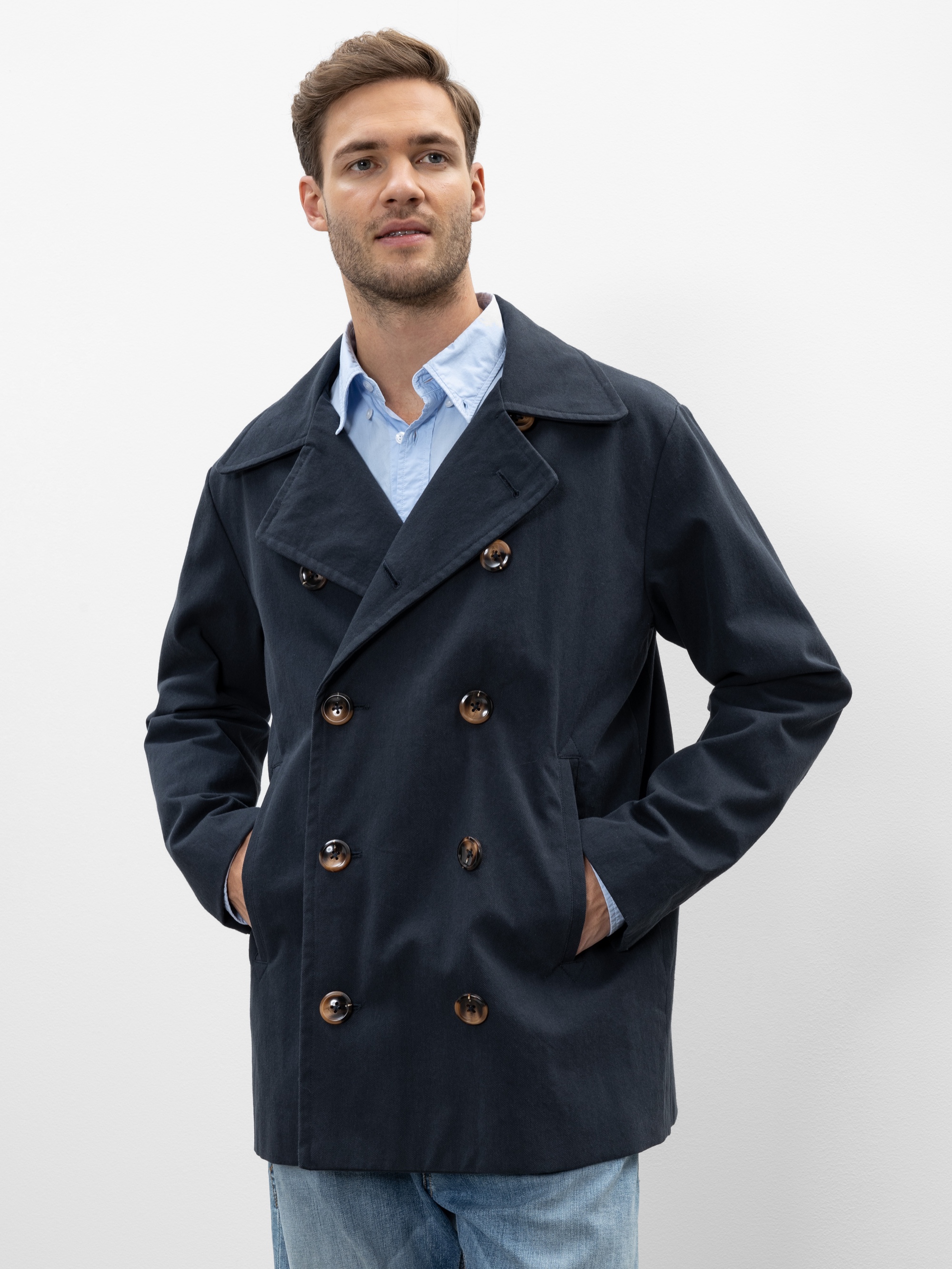 Ein Mann, der einen marineblauen zweireihigen Mantel über einem hellblauen Hemd trägt, steht mit den Händen in den Taschen. Er hat kurzes braunes Haar und einen gestutzten Bart. Er blickt leicht zur Seite und lächelt.