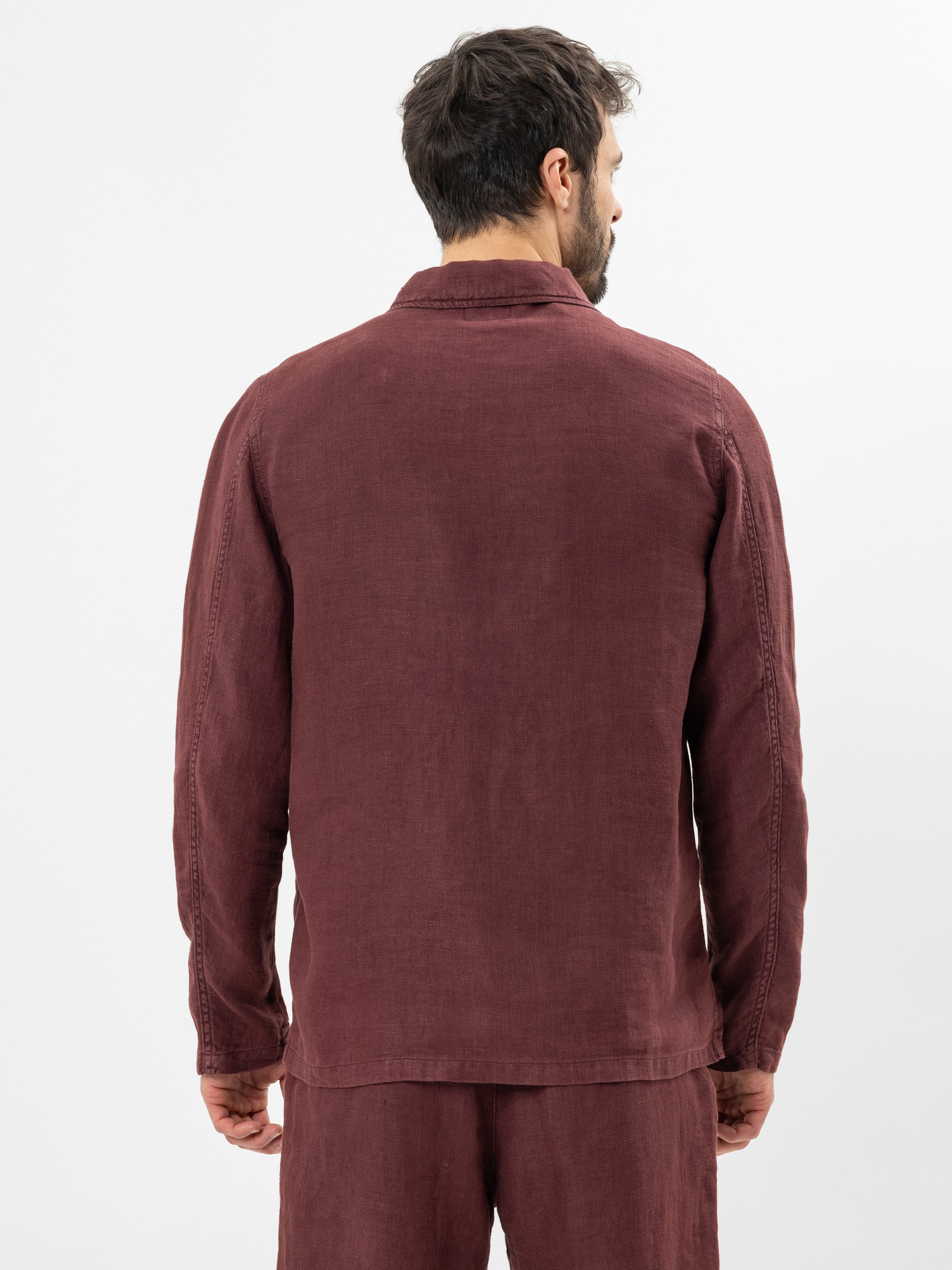 Ein Mann mit kurzen dunklen Haaren wird von hinten gezeigt. Er trägt ein langärmliges, kastanienbraunes Leinenhemd und eine passende Hose. Er steht vor einem schlichten weißen Hintergrund.