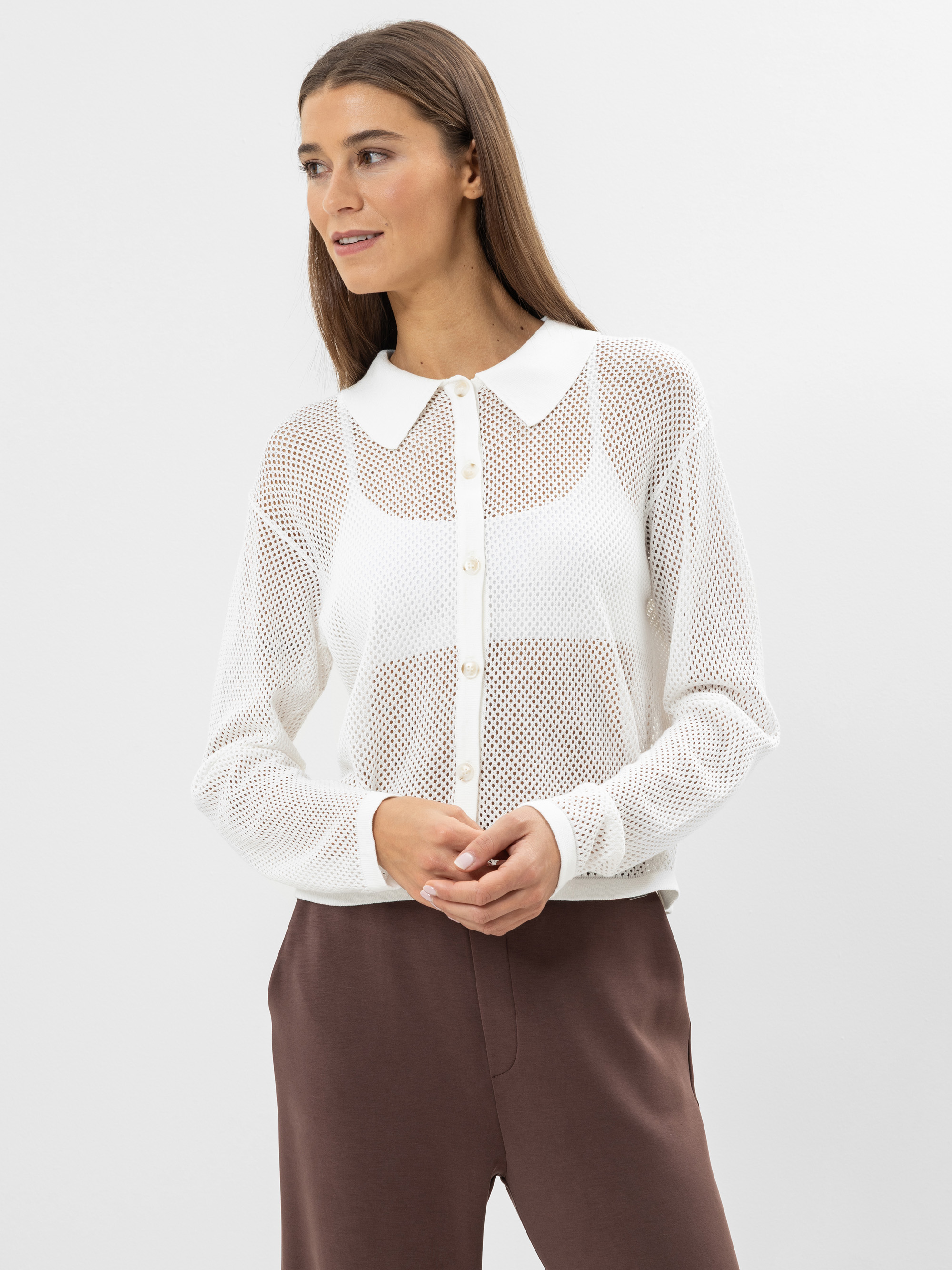 Eine Frau, die ein weißes, langärmeliges, durchsichtiges Netzhemd über einem weißen Oberteil und eine braune Hose mit hoher Taille trägt, steht vor einem schlichten weißen Hintergrund, schaut zur Seite und lächelt leicht.