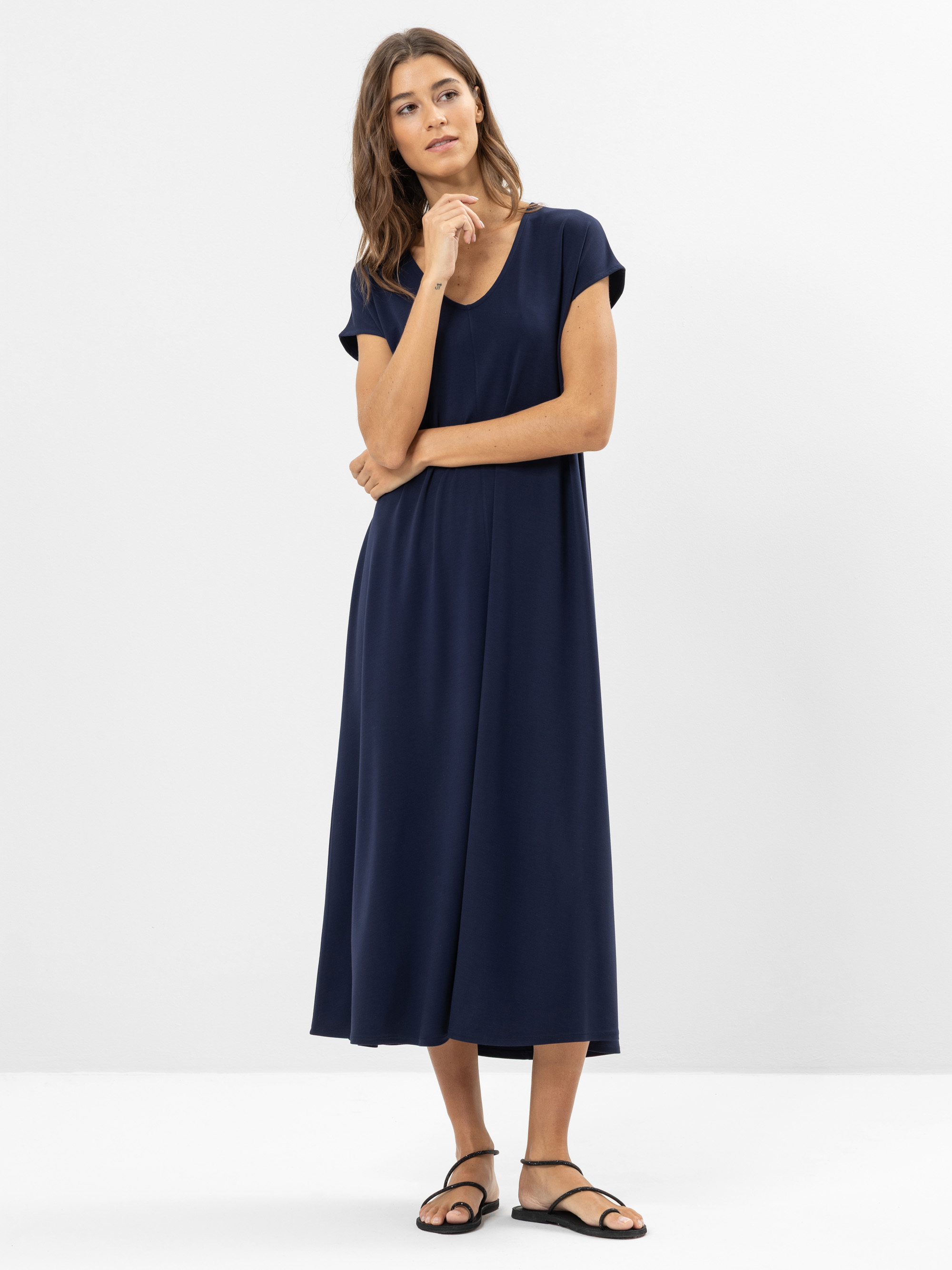 Eine Frau steht vor einem schlichten weißen Hintergrund, trägt ein marineblaues, kurzärmeliges, knöchellanges Kleid und schwarze flache Sandalen. Sie hat langes braunes Haar und blickt leicht zur Seite, eine Hand ruht auf ihrem Kinn.