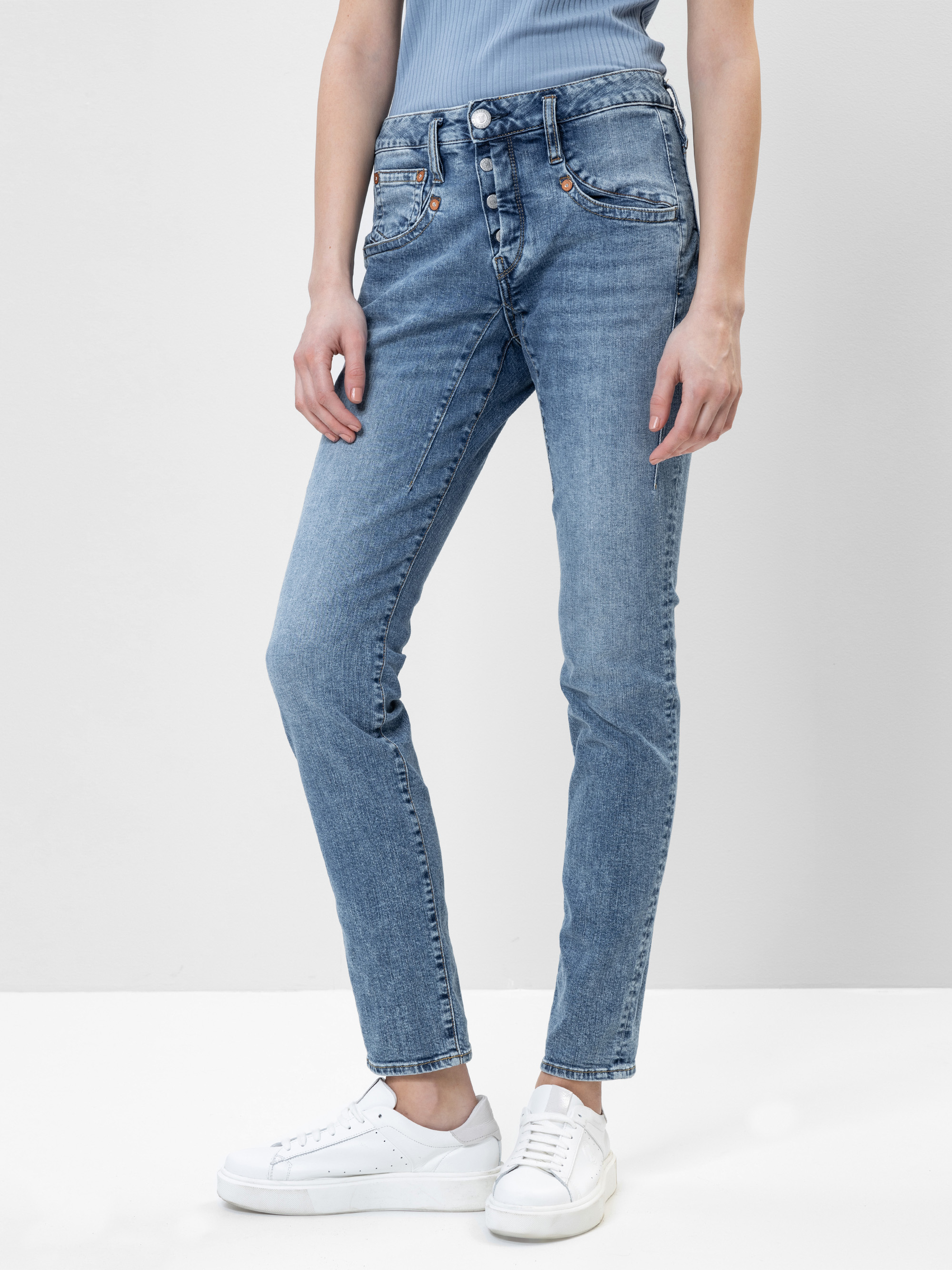 Eine Person, die eine hellblaue Röhrenjeans, ein hellblaues geripptes Oberteil und weiße Turnschuhe trägt, steht vor einem schlichten hellen Hintergrund. Nur die untere Hälfte ihres Körpers ist sichtbar.