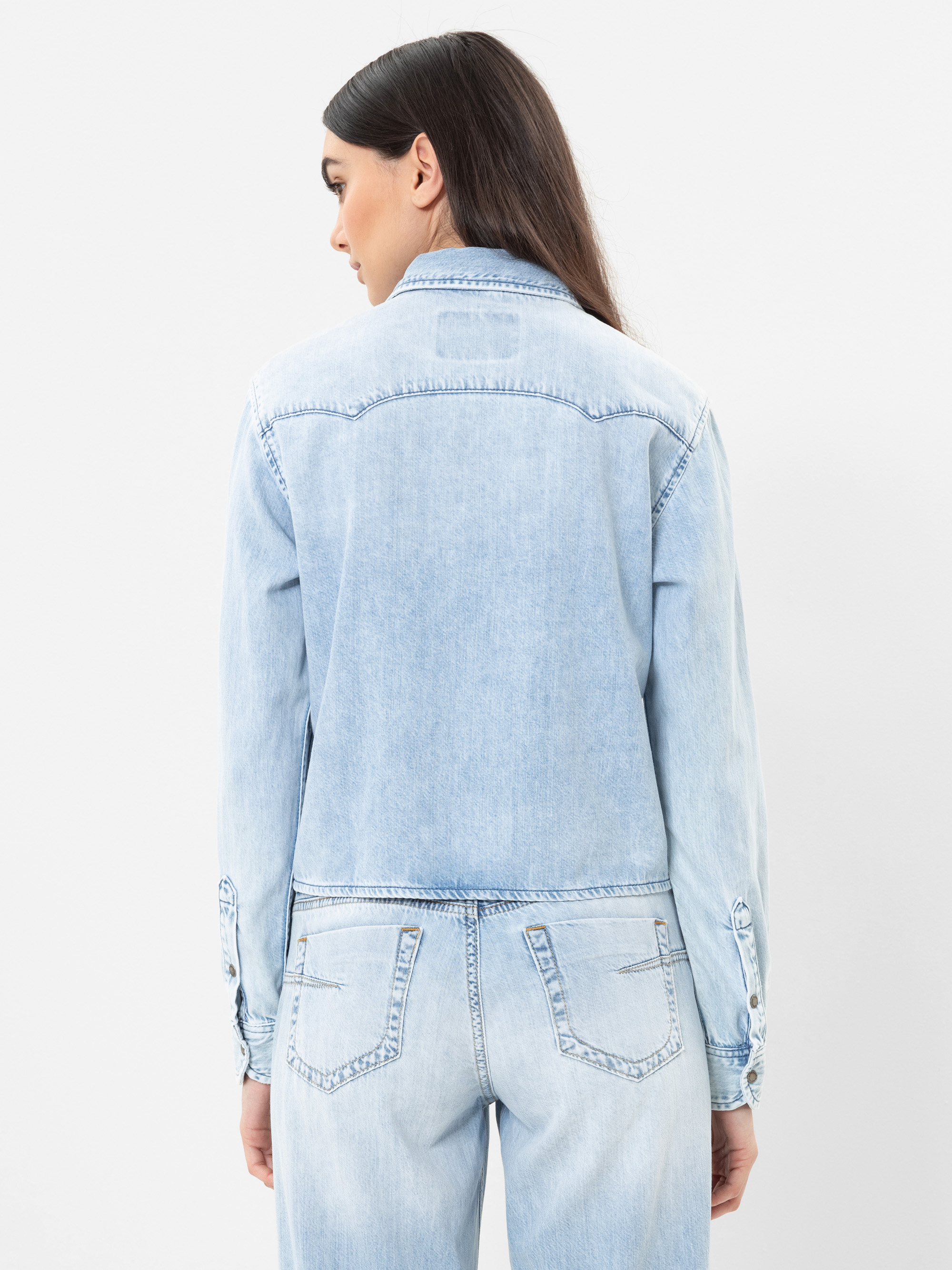 Eine Frau mit langen dunklen Haaren trägt eine hellblaue Jeansjacke und dazu passende Jeans, die von hinten vor einem schlichten weißen Hintergrund gezeigt werden.