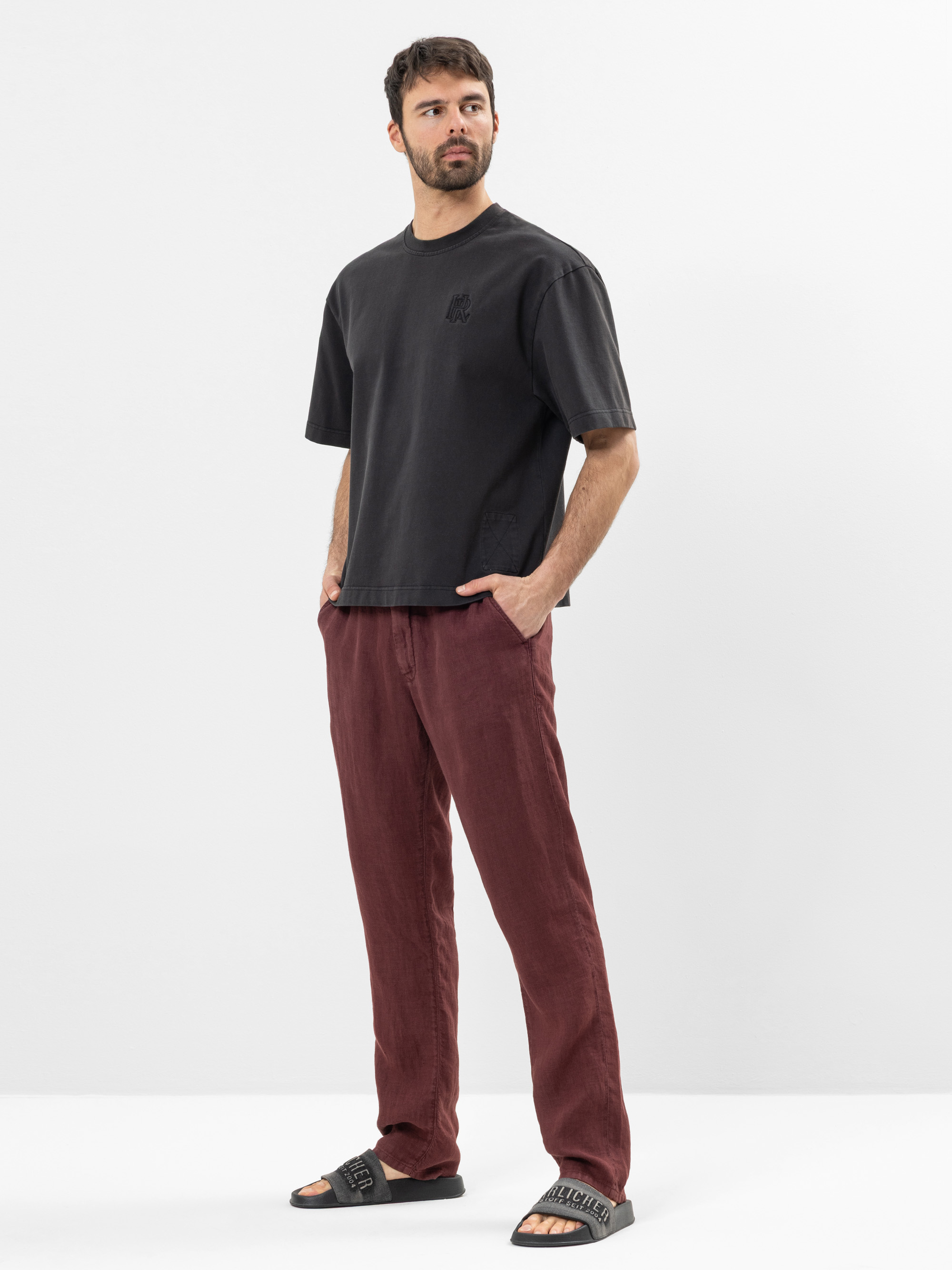 Ein Mann steht vor einem schlichten weißen Hintergrund und trägt ein lockeres schwarzes T-Shirt, eine kastanienbraune Hose und schwarze Slide-Sandalen. Seine Hände stecken in den Taschen, und er schaut leicht zur Seite.