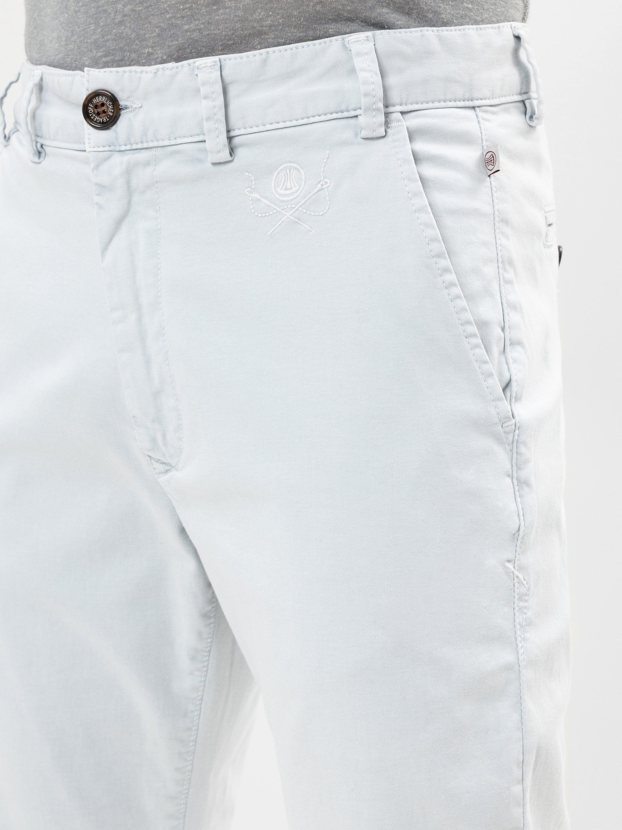 Nahaufnahme einer Person, die eine hellgraue Hose mit Knopf und Reißverschluss, Seitentaschen, Gürtelschlaufen und einem dezenten gestickten Logo über der linken Tasche trägt. Die Person trägt außerdem ein graues Hemd, das in die Hose gesteckt ist.