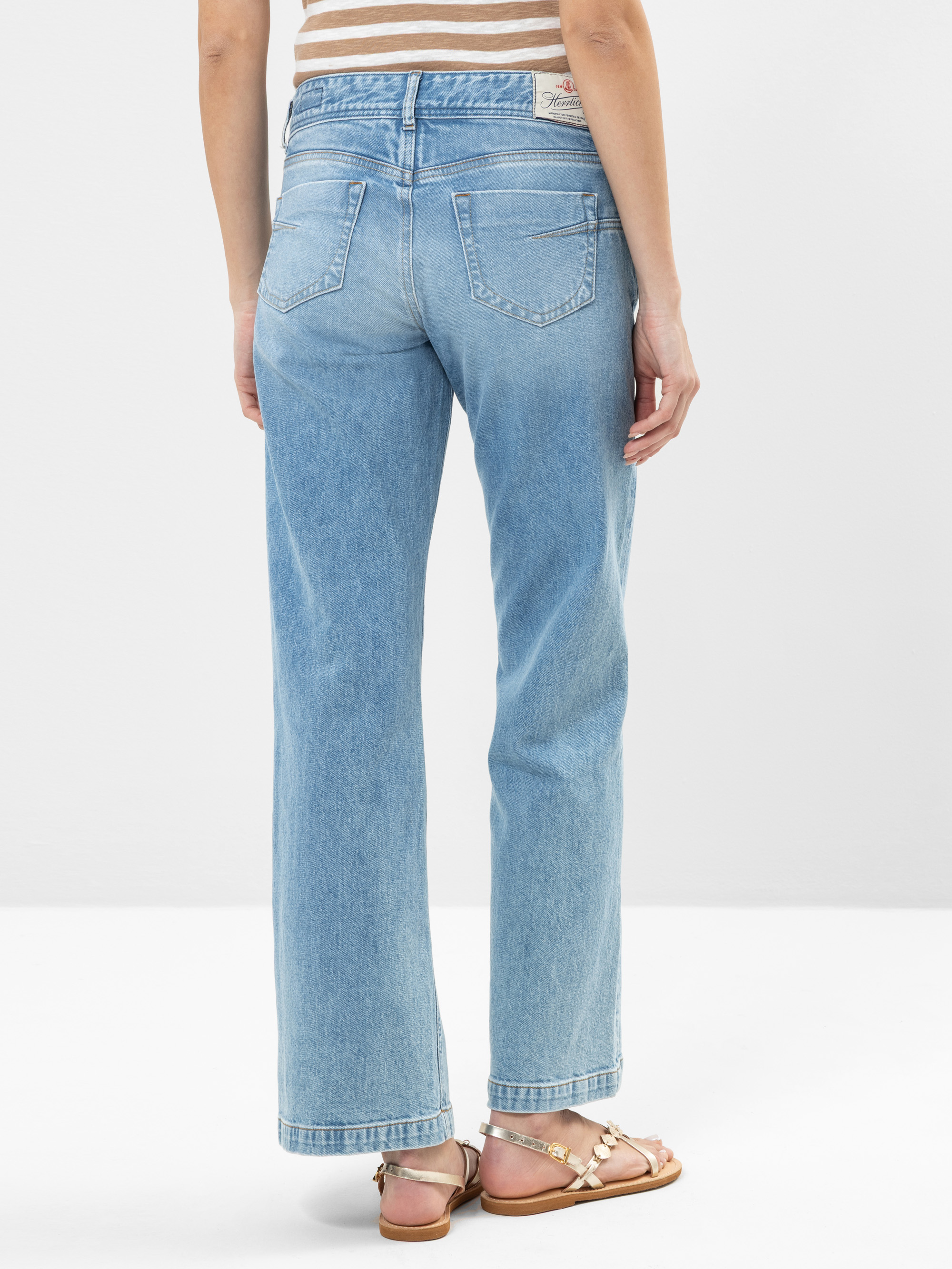 Eine Person, die eine hellblaue, hochgekrempelte Jeans und hellbraune Sandalen trägt, steht mit dem Gesicht zur Seite. Die Jeans hat Gesäßtaschen und einen Lederaufnäher am Bund. Der Hintergrund ist einfarbig und hell gefärbt.