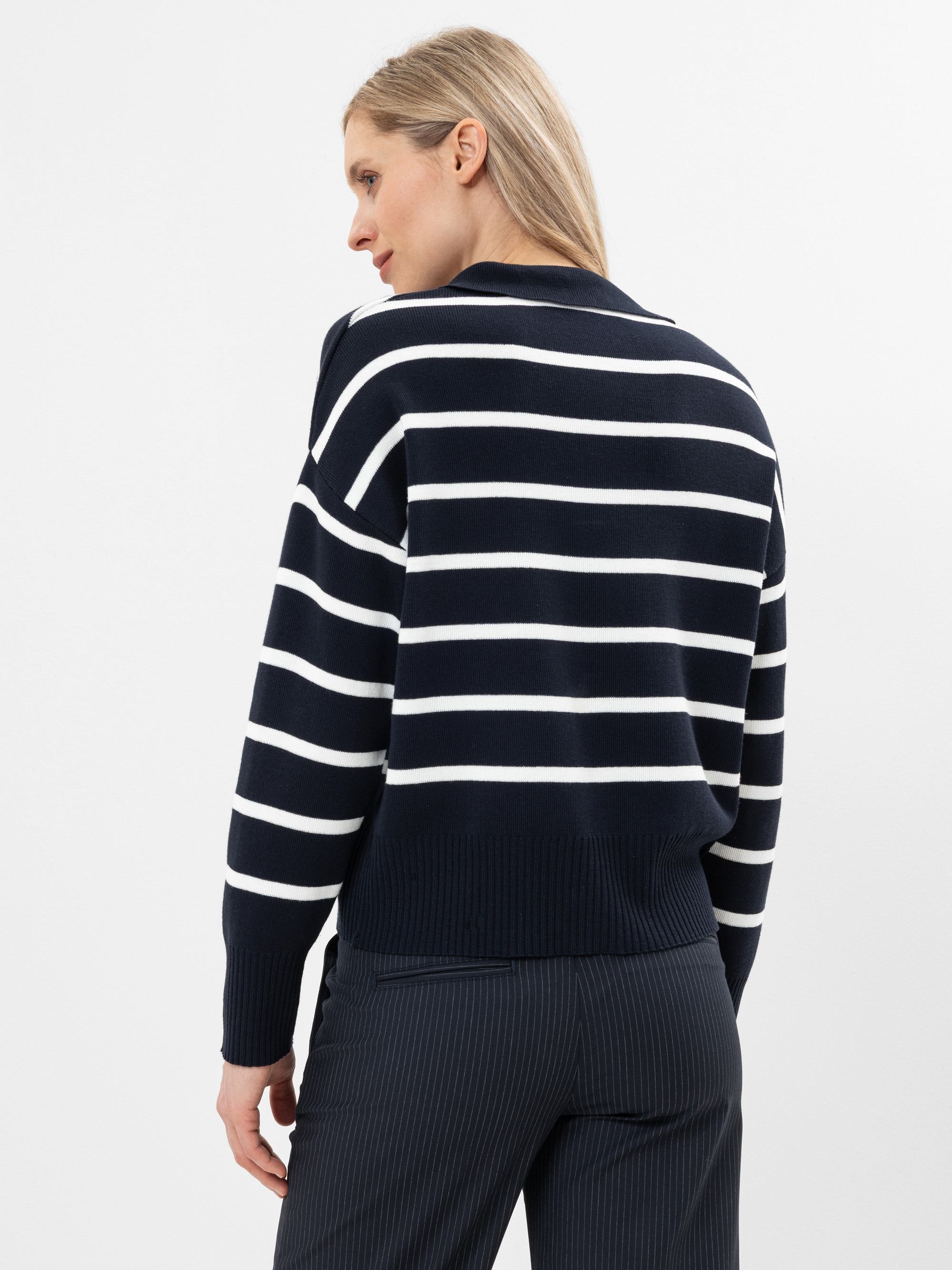 Eine Frau steht mit dem Gesicht zur Wand und trägt einen marineblauen Pullover mit weißen Querstreifen und eine dunkle Hose mit Nadelstreifen. Der Hintergrund ist einfarbig weiß.