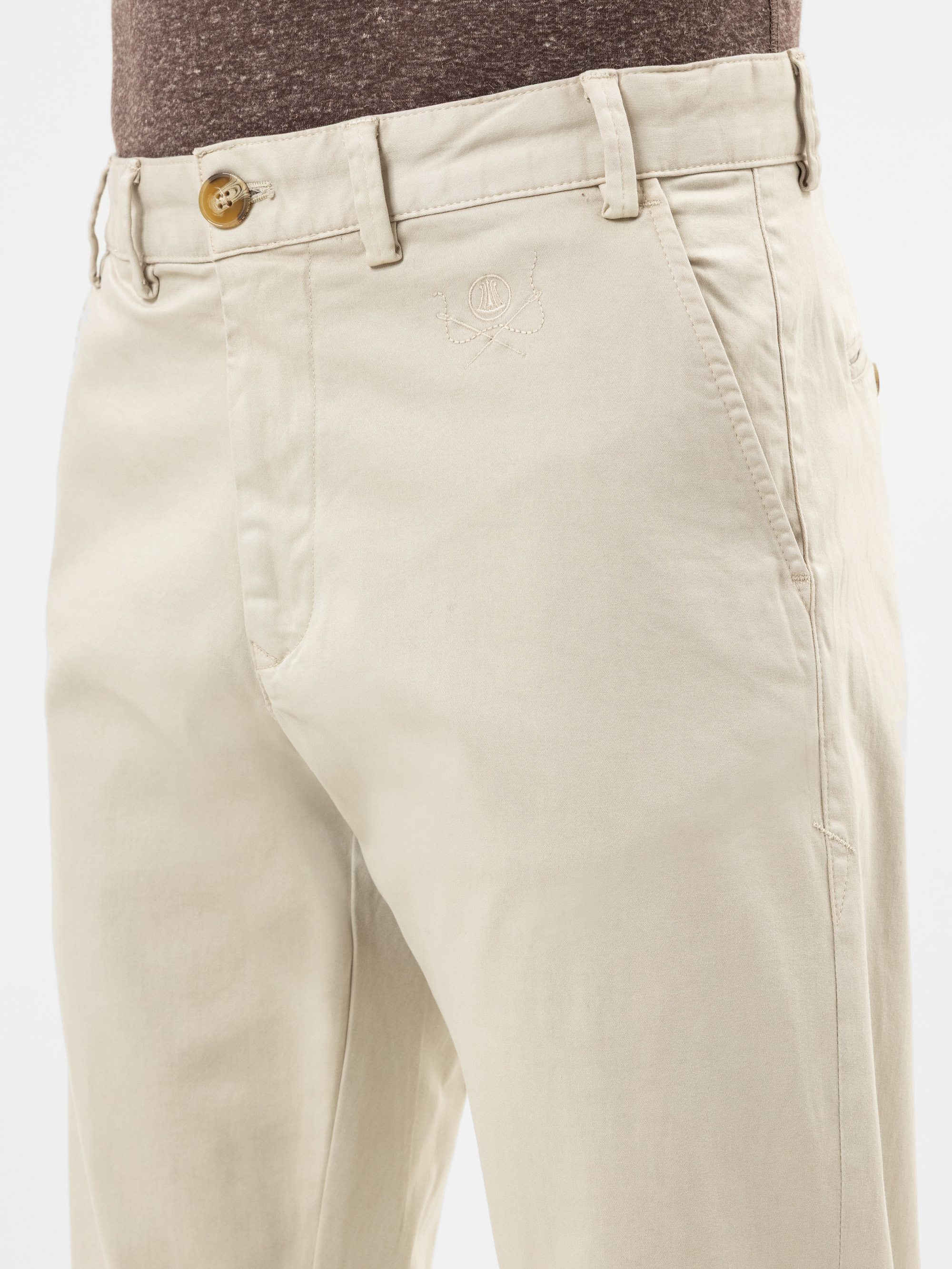 Nahaufnahme einer Person, die eine beigefarbene Chino-Hose mit Knopf und Reißverschluss, Gürtelschlaufen und einem gestickten Wappen auf der Tasche trägt, kombiniert mit einem braunen Hemd.