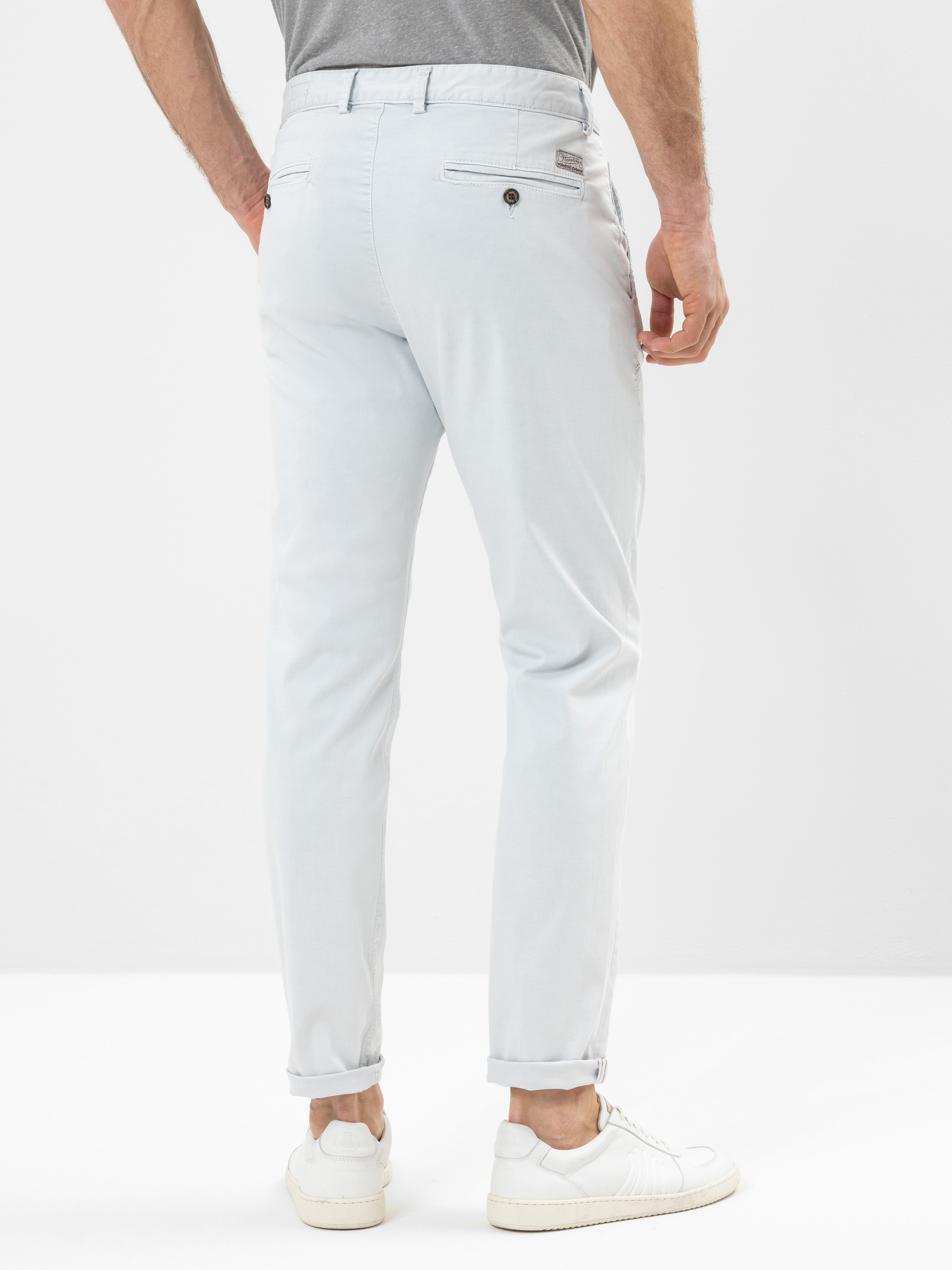 Ein Mann, der eine hellgraue Hose, ein graues Hemd und weiße Turnschuhe trägt, steht mit dem Gesicht zum Boden, die Hände an den Seiten. Die Hose ist an den Knöcheln mit Manschetten geschlossen.