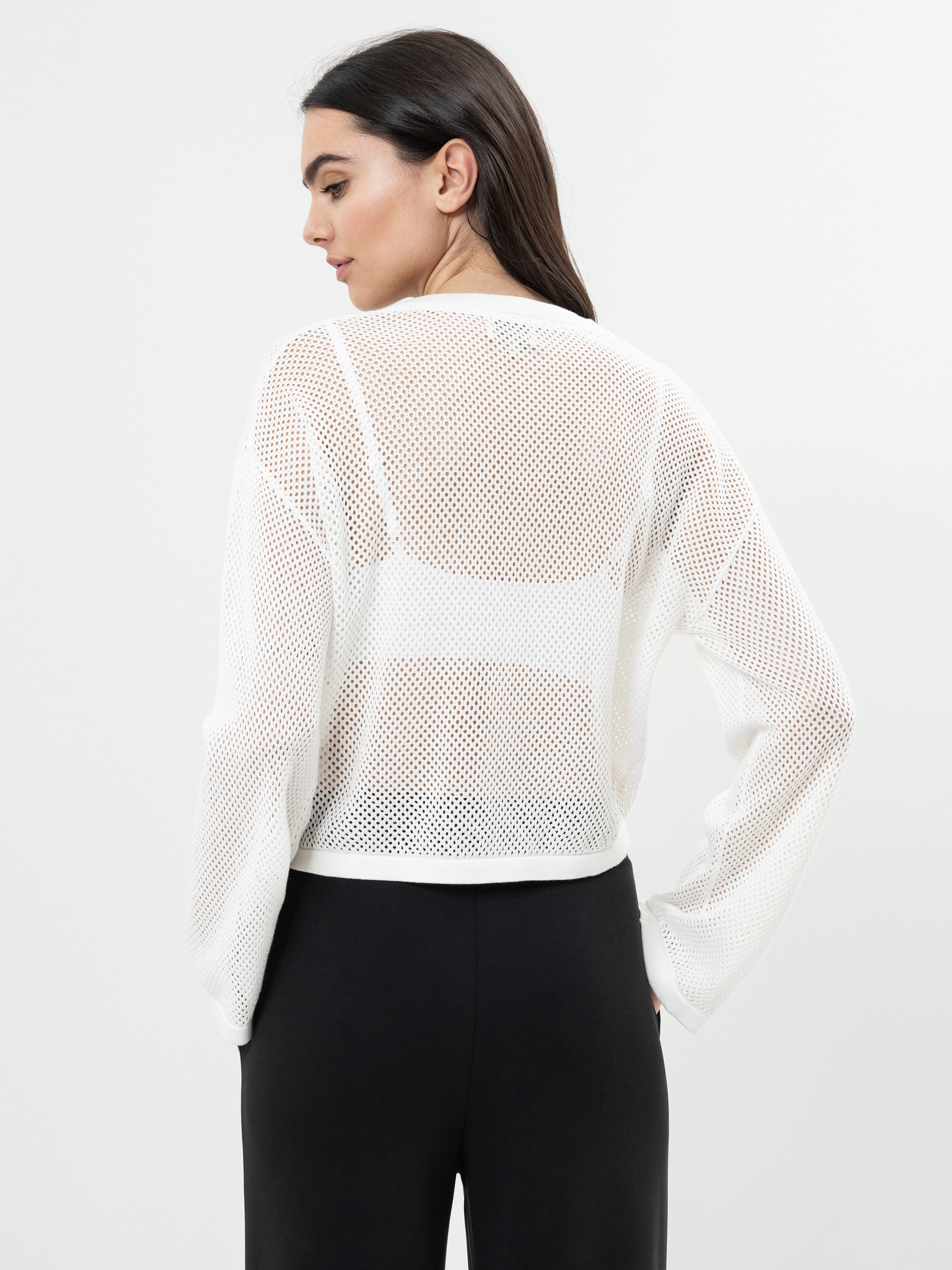 Frau mit langen dunklen Haaren, die ein durchsichtiges weißes Mesh-Top über einem weißen Bralette und eine schwarze Hose mit hoher Taille trägt und mit dem Rücken zur Kamera vor einem schlichten Hintergrund steht.