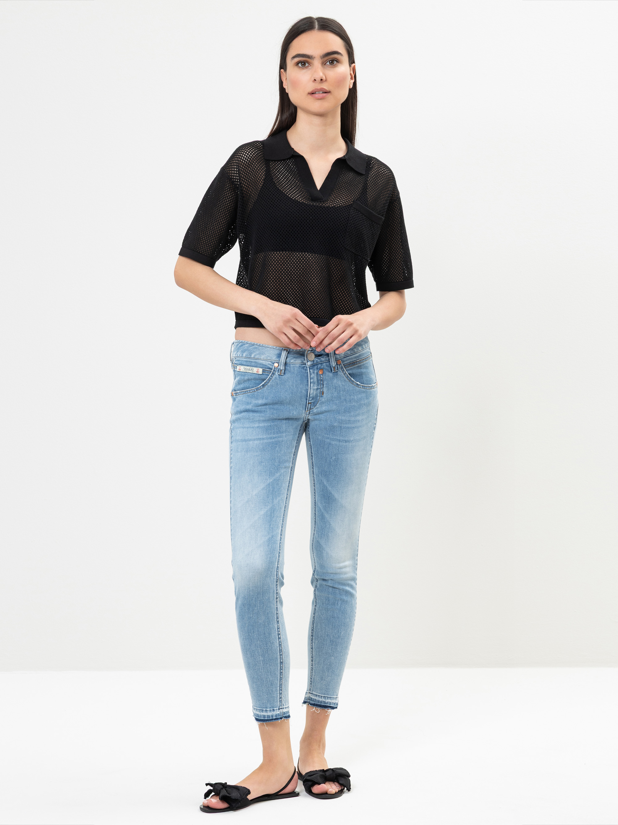 Eine Frau steht vor einem schlichten weißen Hintergrund und trägt ein schwarzes kurzärmeliges Netzoberteil über einem schwarzen Bralette, hellblaue Röhrenjeans und schwarze Slipper mit Schleifen. Sie hat glattes dunkles Haar und einen neutralen Gesic
