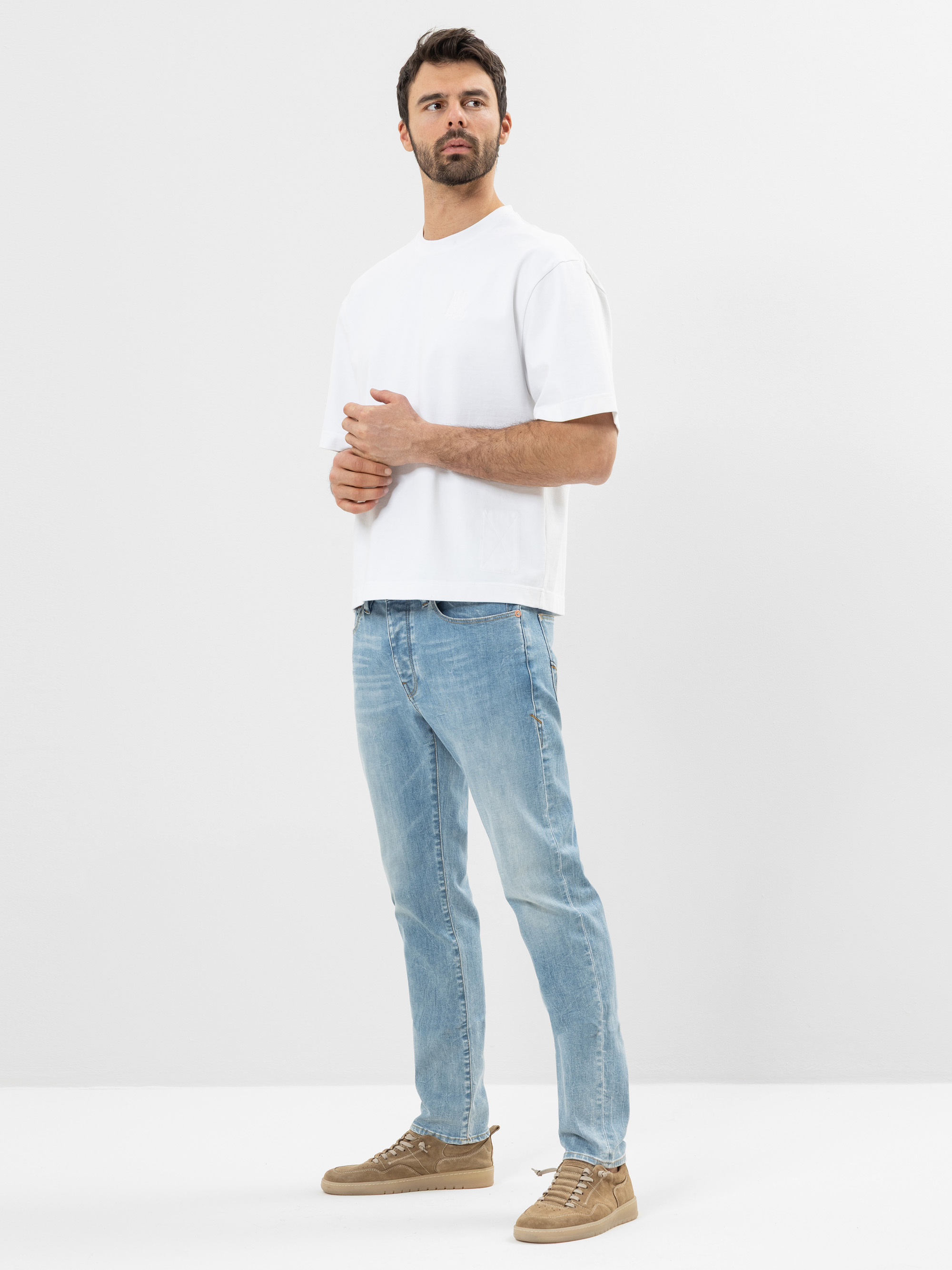 Ein Mann steht in einem schlichten weißen Raum und trägt ein lockeres weißes T-Shirt, hellblaue Jeans und hellbraune Turnschuhe. Er schaut leicht zur Seite und stützt eine Hand auf sein anderes Handgelenk.