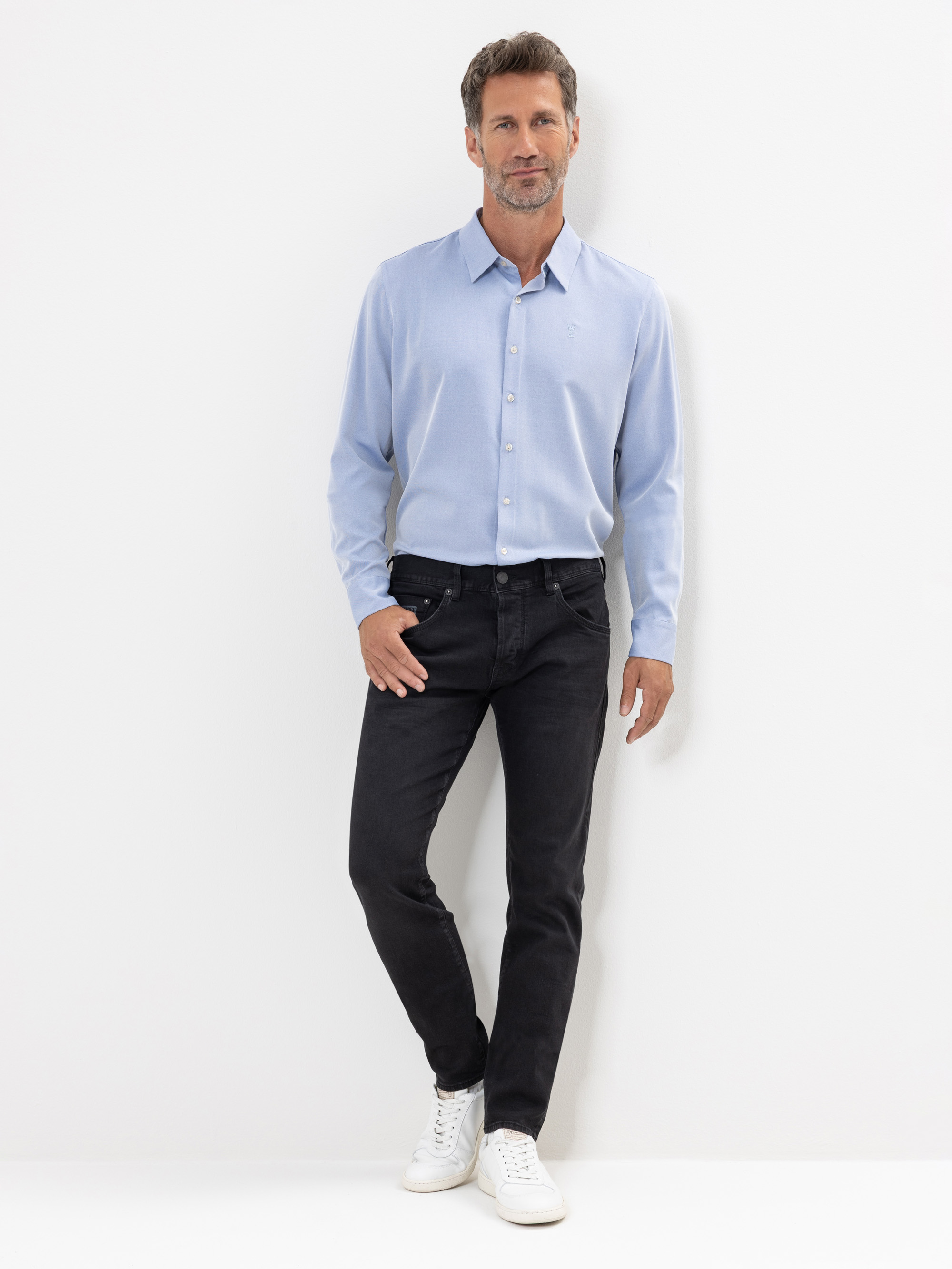 Ein Mann steht vor einem weißen Hintergrund und trägt ein hellblaues Button-up-Hemd, schwarze Jeans und weiße Turnschuhe. Er hat kurze Haare und einen Bart und schaut mit entspannter Haltung direkt in die Kamera.