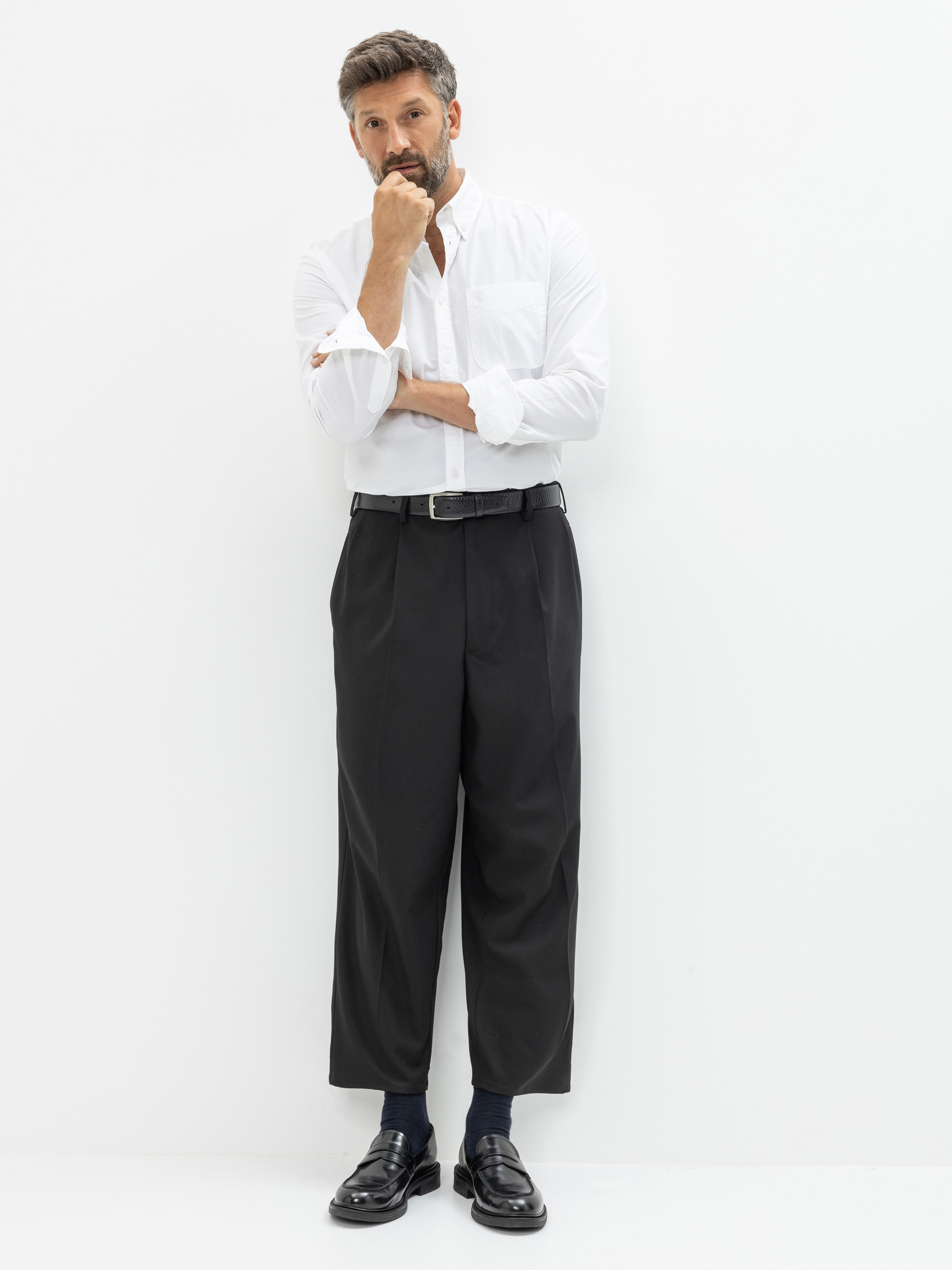 Ein Mann steht vor einem weißen Hintergrund. Er trägt ein weißes langärmeliges Hemd, das in eine schwarze Hose mit weitem Bein und Gürtel gesteckt ist, dunkle Socken und schwarze Halbschuhe. Er stützt eine Hand auf sein Kinn und blickt nachdenklich