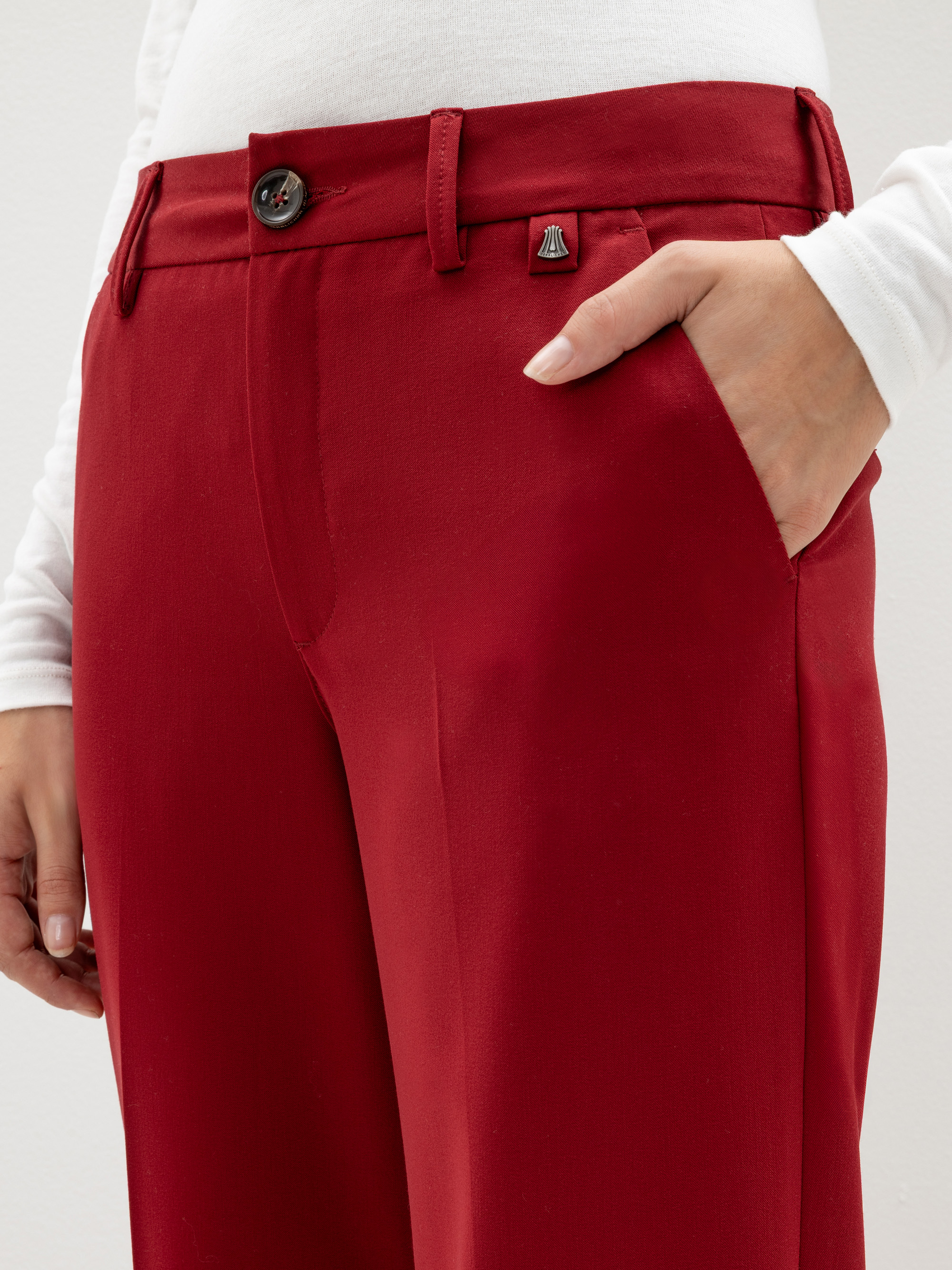Eine Person, die eine hochgeschnittene, rote Hose mit einem Knopf und einem kleinen Metallemblem in der Nähe des Hosenbundes und ein weißes, langärmeliges Oberteil trägt; eine Hand steckt in der rechten Tasche.