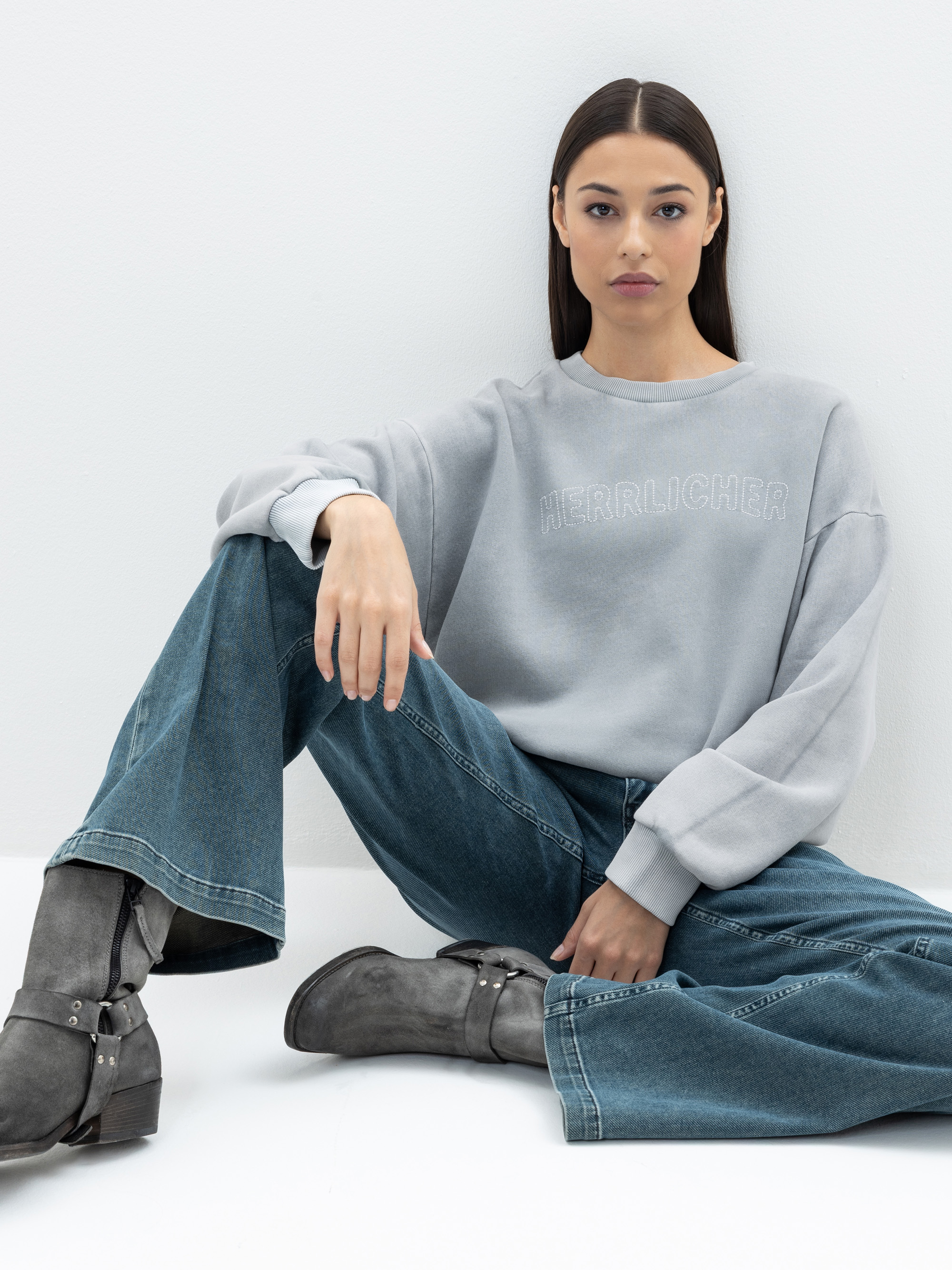Eine Frau mit langen dunklen Haaren sitzt auf dem Boden vor einer weißen Wand. Sie trägt ein hellgraues Sweatshirt mit der Aufschrift HER*RLICHER auf der Vorderseite, blaue Jeans und graue Stiefel. Ihr Gesichtsausdruck ist neutral.