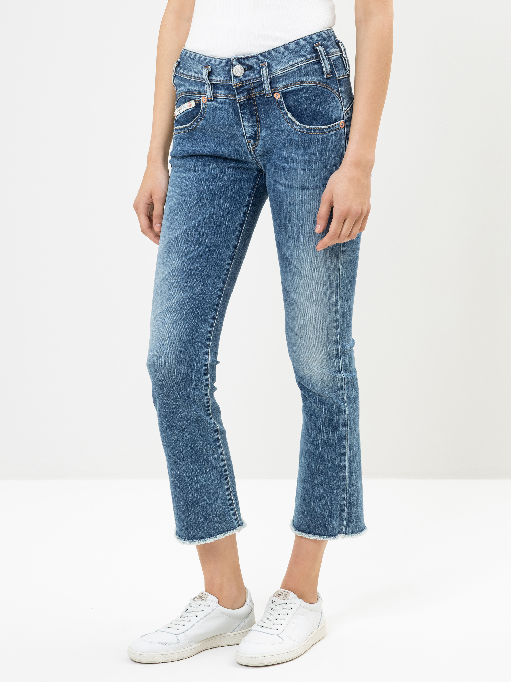 Eine Person, die blaue Jeans mit ausgefransten Säumen, ein weißes geripptes Oberteil und weiße Turnschuhe trägt, steht vor einem schlichten weißen Hintergrund.