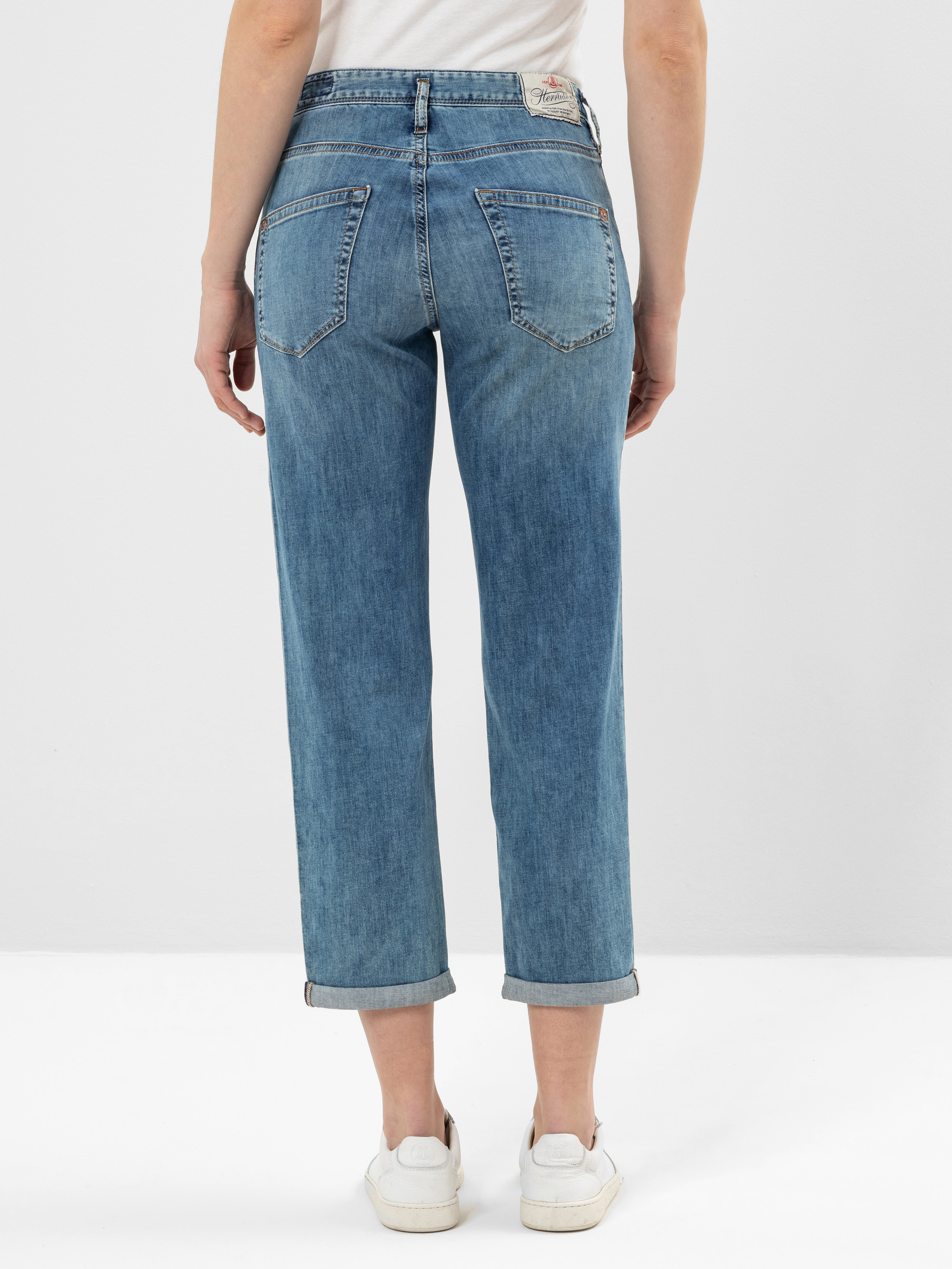 Eine Person, die eine hellblaue Jeans mit geradem Bein und weiße Turnschuhe trägt, steht mit dem Rücken zur Kamera vor einem schlichten, hellen Hintergrund.