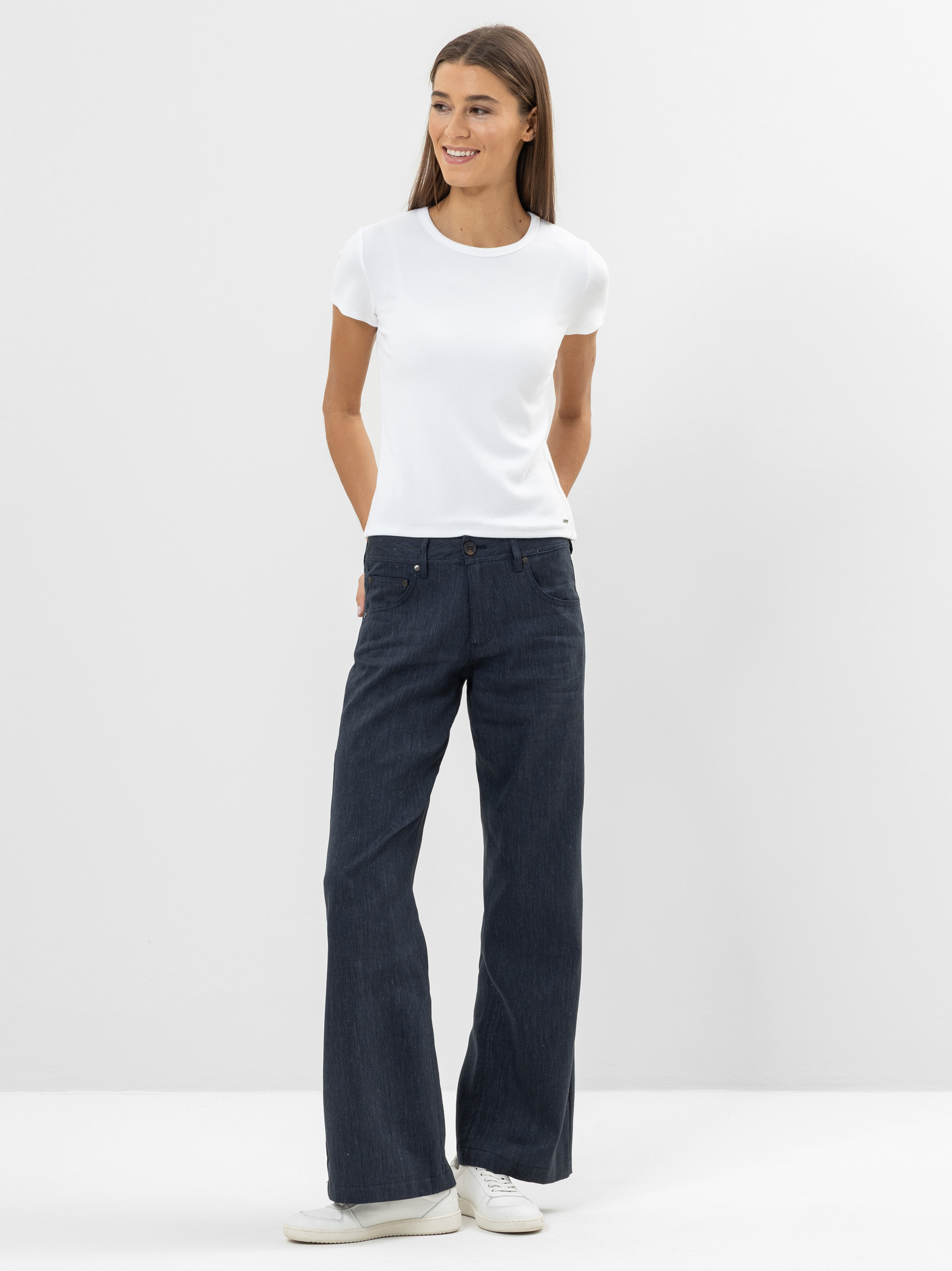 Eine Person steht vor einem schlichten weißen Hintergrund, trägt ein weißes, kurzärmeliges T-Shirt, dunkle, weit geschnittene Jeans und weiße Turnschuhe und lächelt mit den Händen auf dem Rücken.