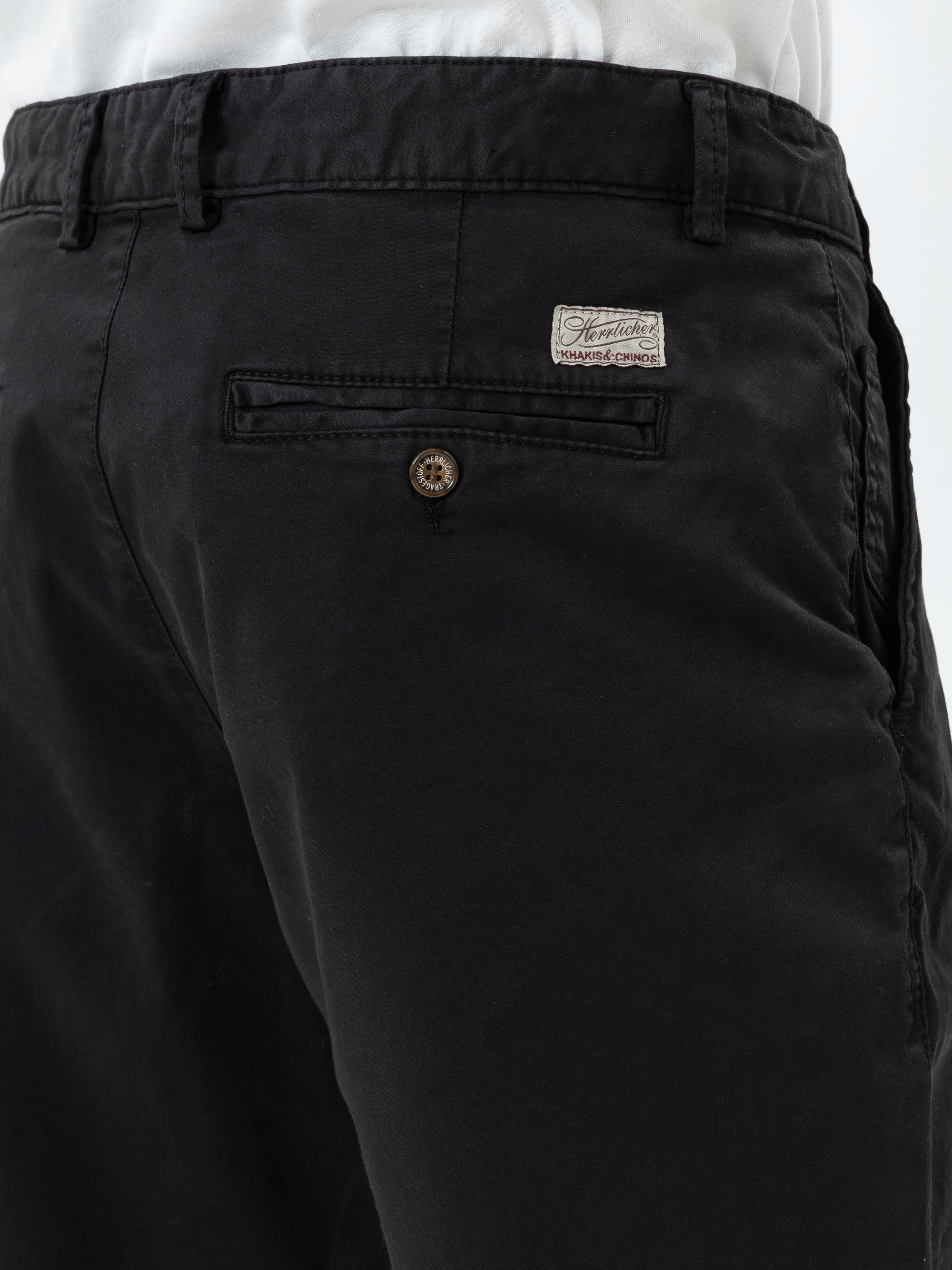 Nahaufnahme einer Person, die eine schwarze Hose mit einer Gesäßtasche, einem braunen Knopf und einem weißen Etikett über der Tasche trägt. Die Person trägt außerdem ein weißes Hemd, das in die Hose gesteckt wird.
