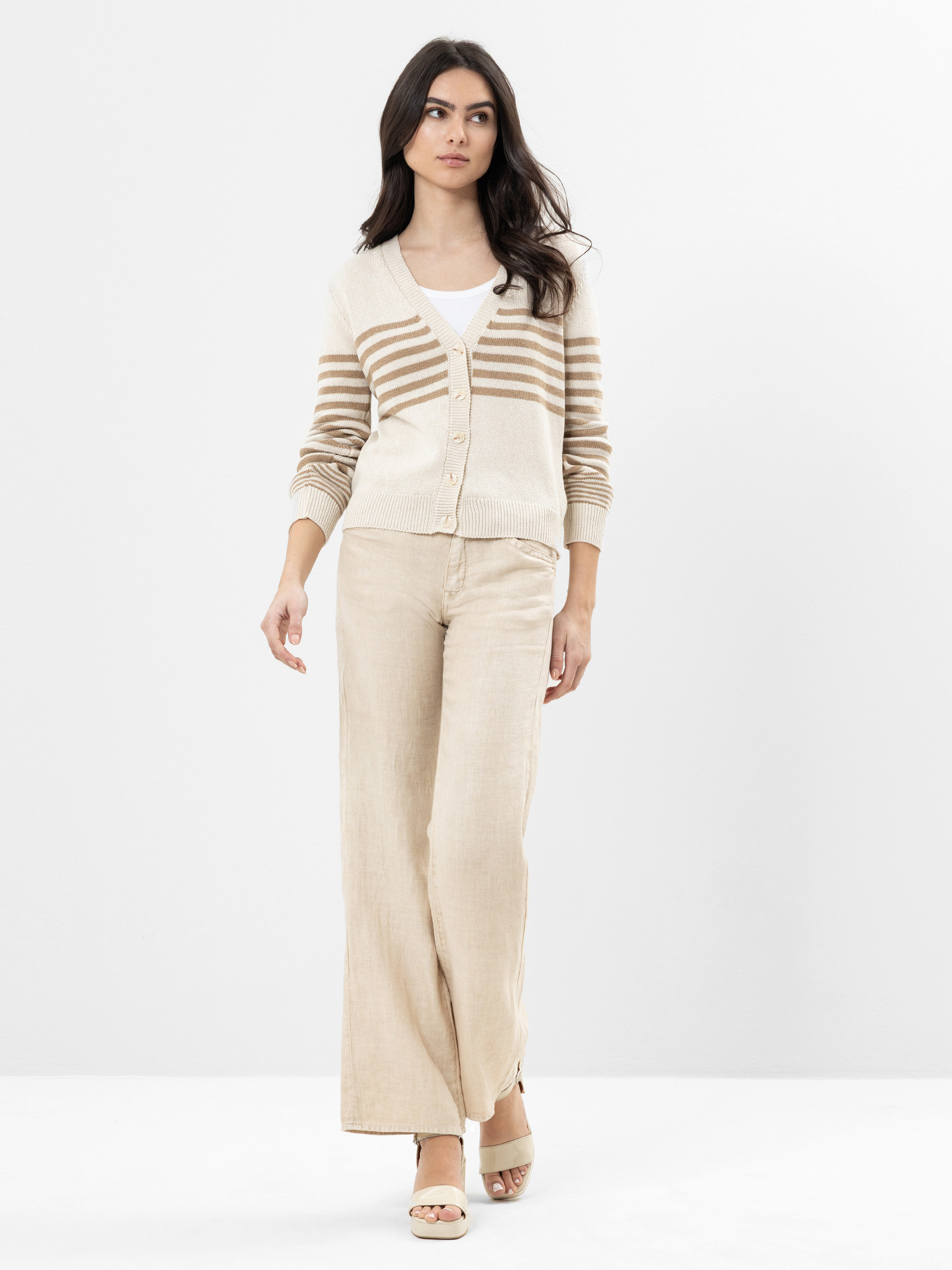 Eine Frau mit langen dunklen Haaren trägt eine creme-beige gestreifte Strickjacke über einem weißen Oberteil, eine beigefarbene Hose mit weitem Bein und beigefarbene Keilsandalen vor einem schlichten weißen Hintergrund.