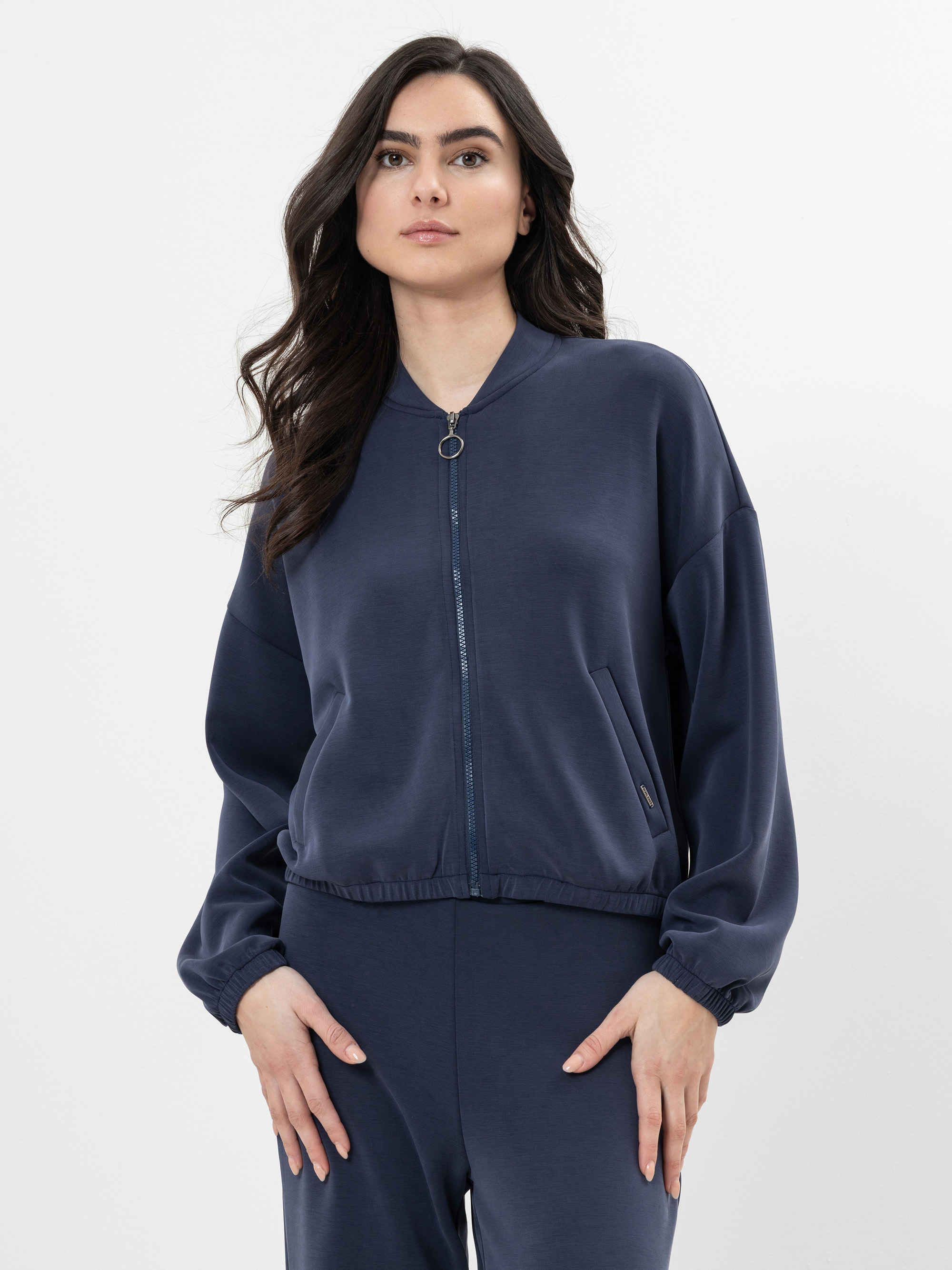 Eine Frau mit langem, dunklem, gewelltem Haar trägt eine marineblaue Jacke mit Reißverschluss und eine dazu passende Hose. Sie steht vor einem schlichten weißen Hintergrund und hat einen neutralen Gesichtsausdruck.