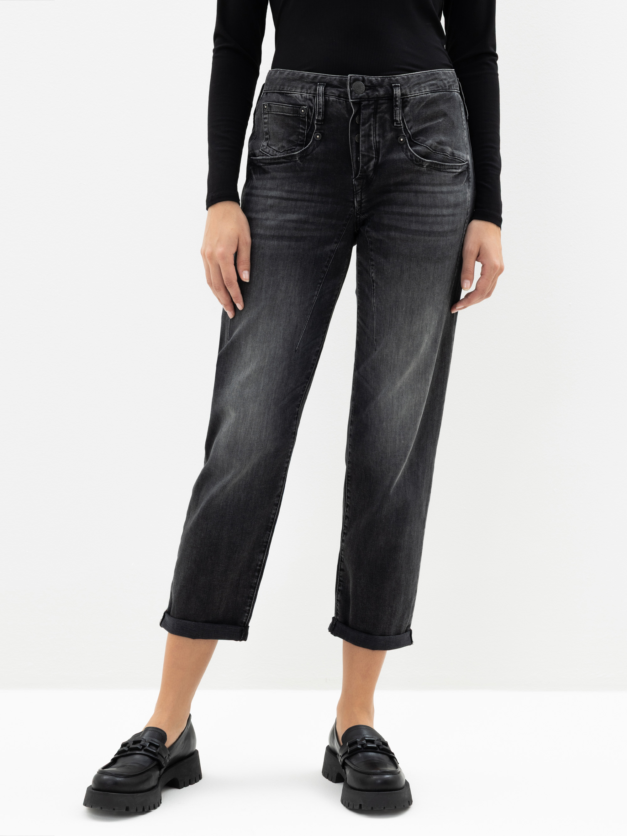 Eine Person, die ein schwarzes langärmeliges Oberteil, verwaschene schwarze Jeans mit aufgerollten Manschetten und schwarze Slipper trägt, steht mit dem Gesicht nach vorne vor einem schlichten weißen Hintergrund.