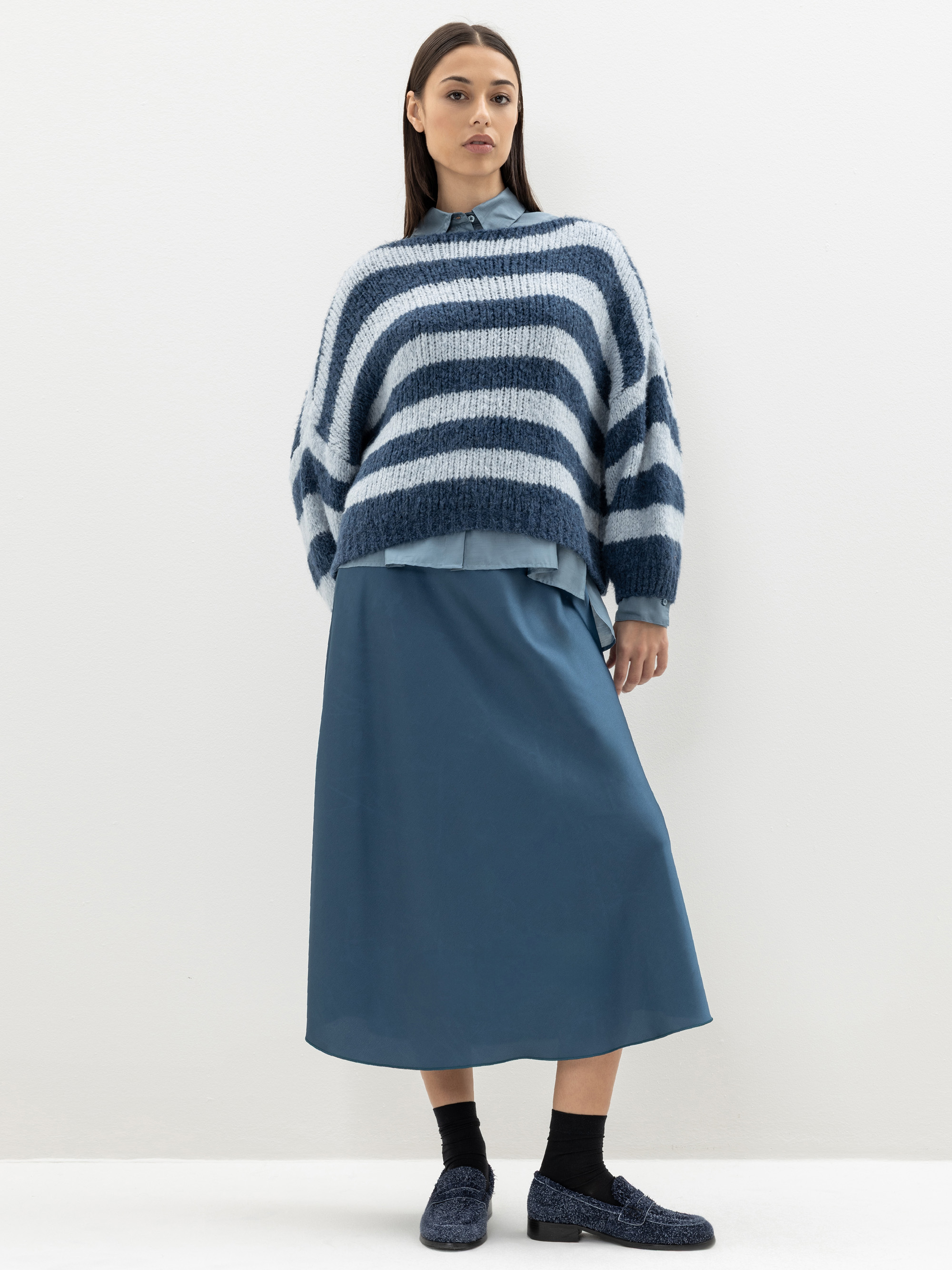 Eine Frau steht vor einer weißen Wand und trägt einen blauen Maxirock, ein hellblaues Button-up-Hemd und einen lockeren, blau-weiß gestreiften Pullover. Dazu trägt sie marineblaue Halbschuhe und schwarze Socken.