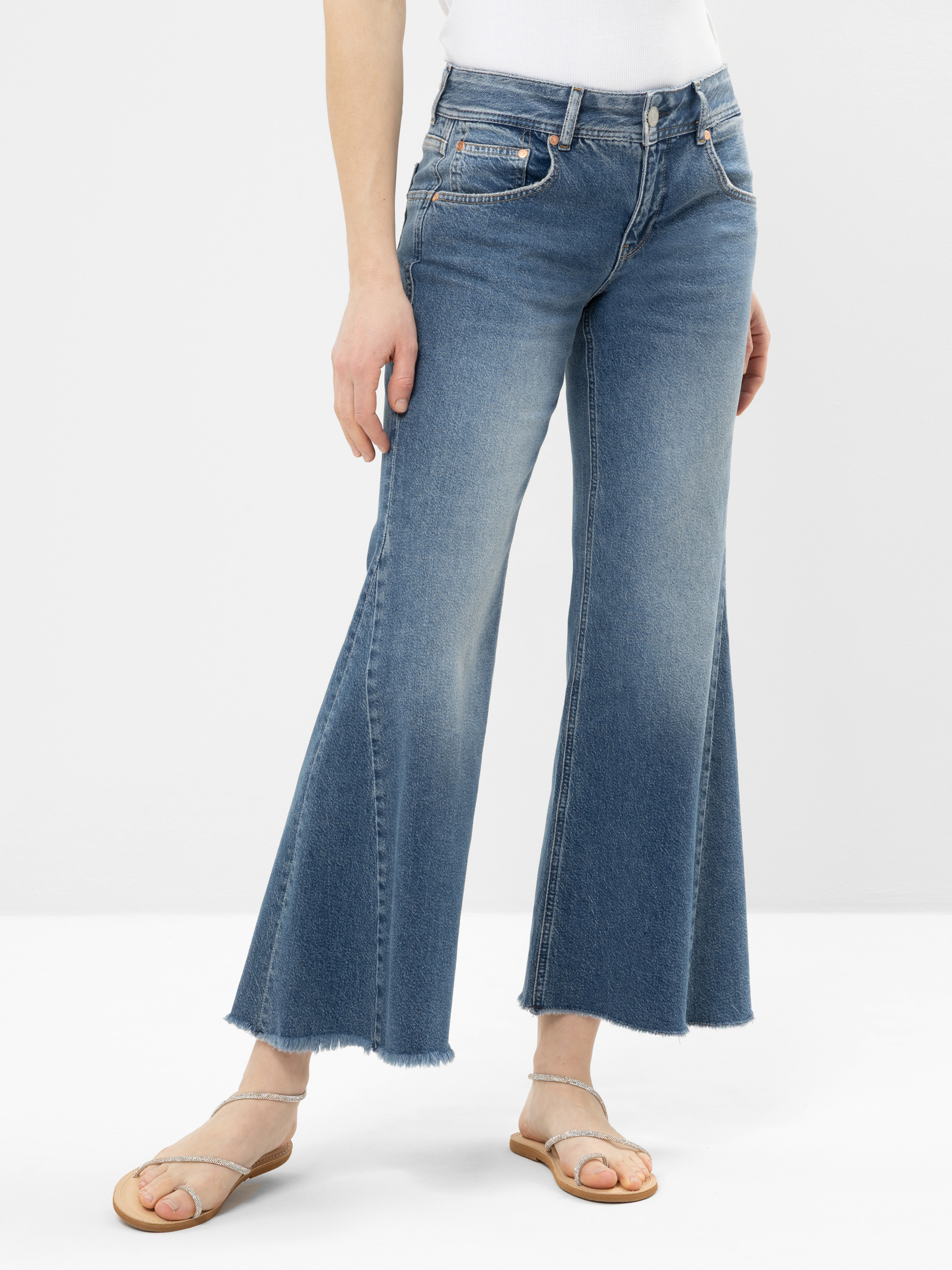 Eine Person, die verblichene blaue Jeans mit weitem Bein und ausgefranstem Saum, ein weißes Oberteil und beigefarbene flache Sandalen trägt, steht vor einem schlichten hellen Hintergrund.