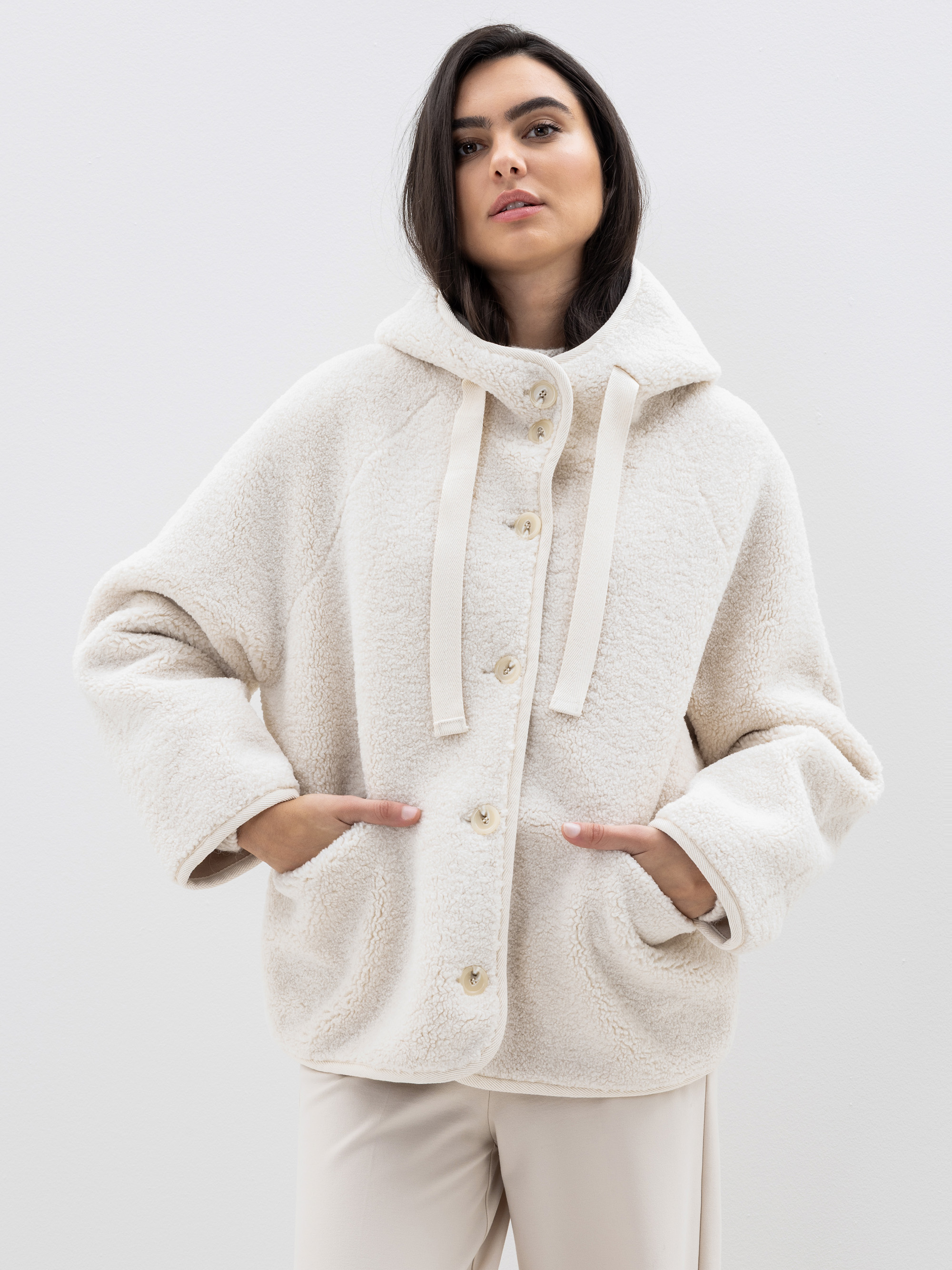 Eine Frau in einer cremefarbenen Fleecejacke mit Kapuze und beigen Kordelzügen, die mit den Händen in den Taschen vor einem schlichten hellen Hintergrund steht.