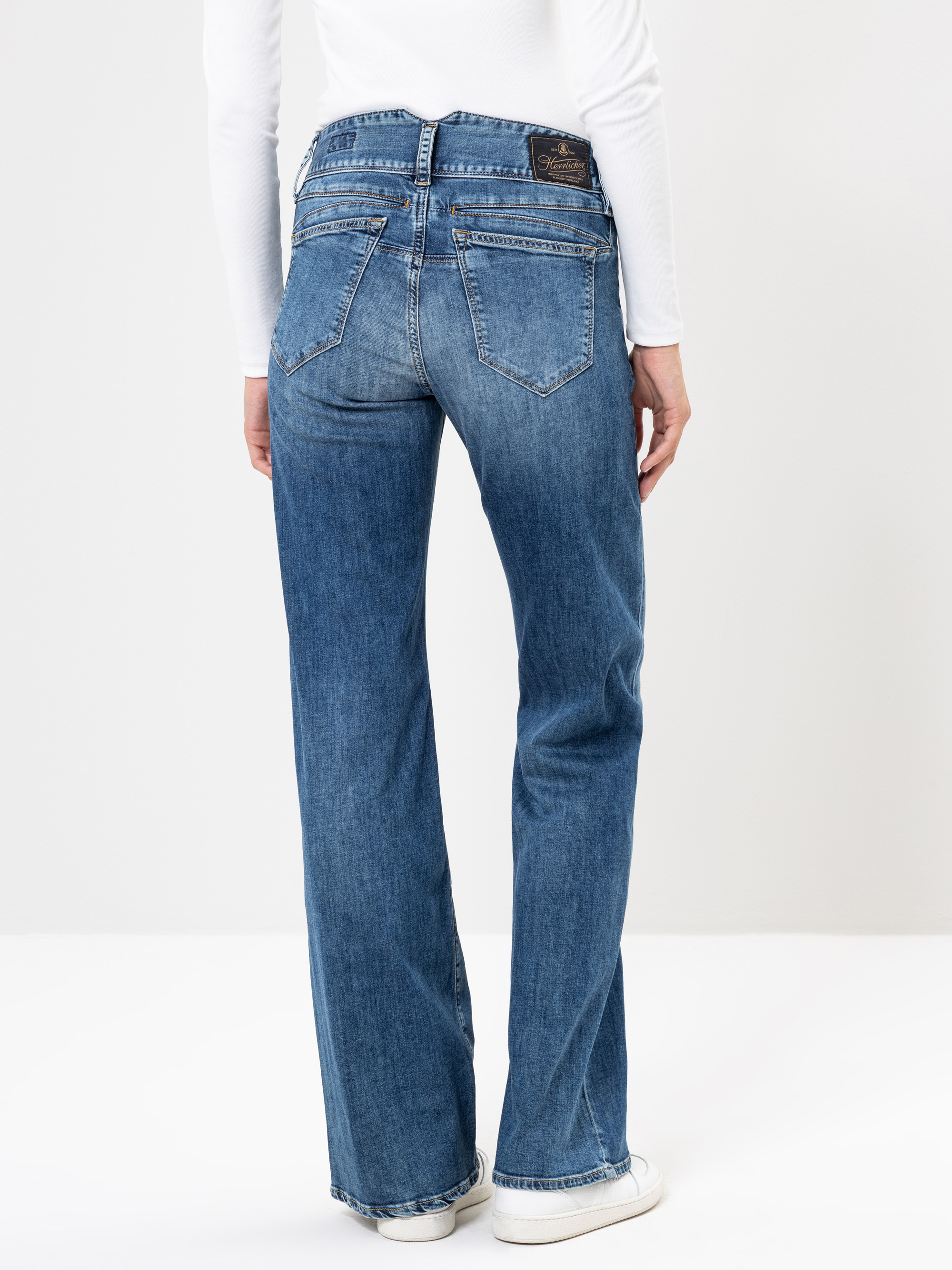 Eine Person, die ein weißes langärmeliges Oberteil, blaue Jeans mit geradem Bein und weiße Turnschuhe trägt, steht mit dem Rücken zur Kamera vor einem schlichten weißen Hintergrund.