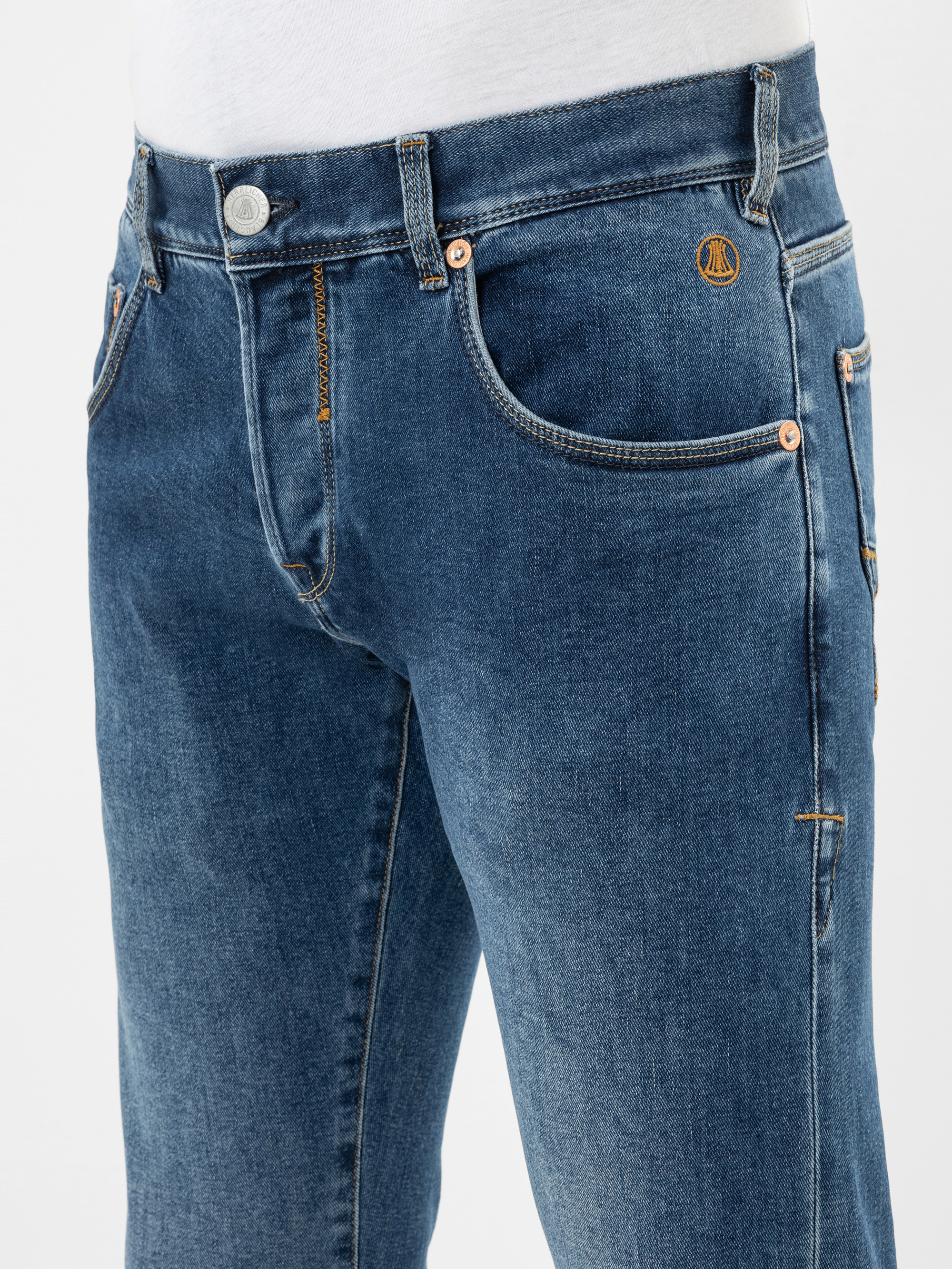 Nahaufnahme einer Person, die eine blaue Jeans mit kupferfarbenen Nieten und einem orangefarbenen gestickten Emblem auf der Vordertasche trägt, dazu ein weißes Hemd.