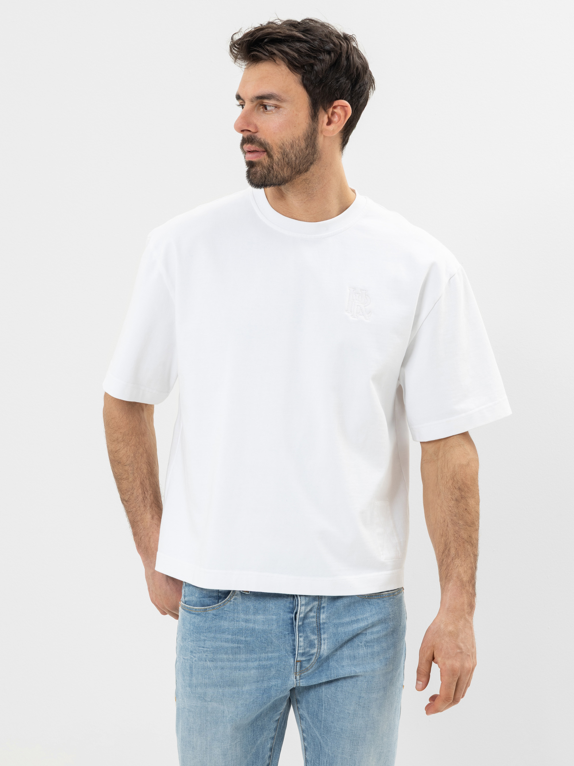 Ein Mann mit dunklen Haaren und einem Bart trägt ein locker sitzendes weißes T-Shirt und hellblaue Jeans. Er steht vor einem schlichten weißen Hintergrund und blickt nach links.